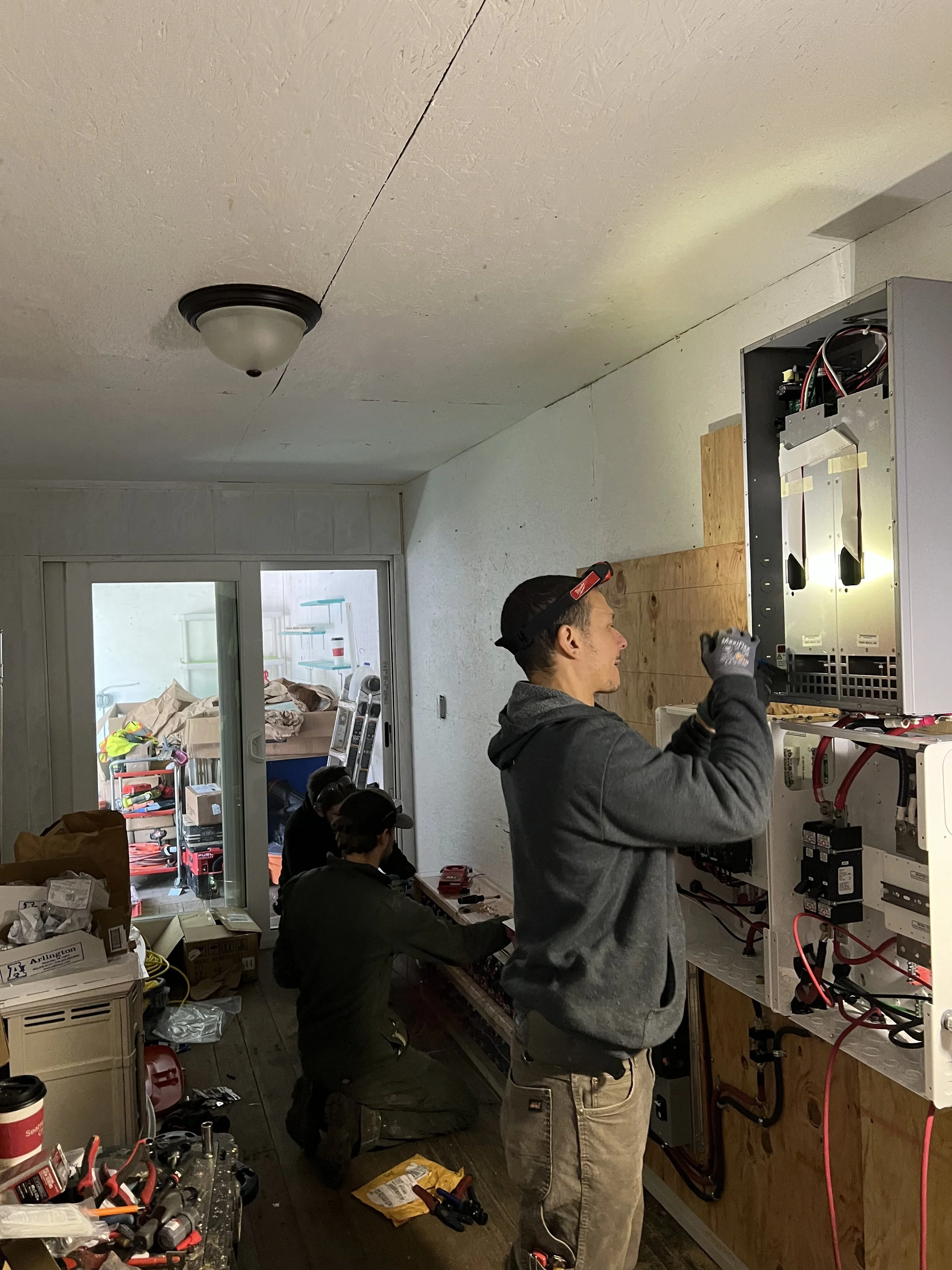 Three men working on electrical or electronic wiring inside a wall panel in a room with cluttered work area.