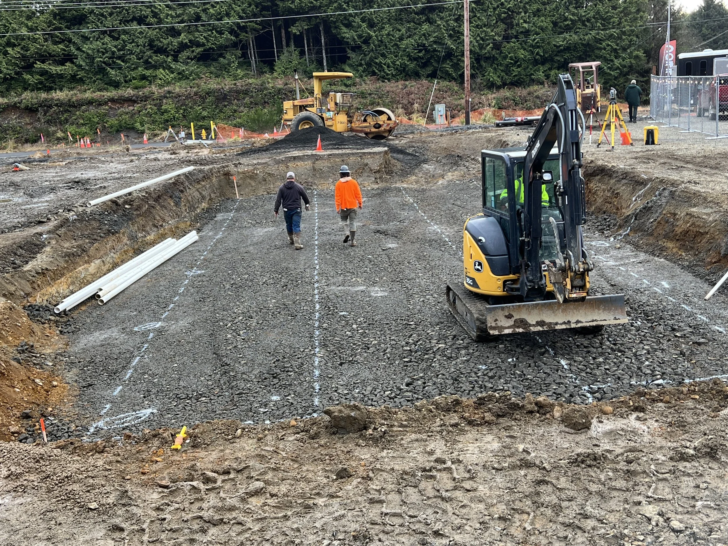 Construction site with roadwork equipment, workers, and trees in the background.