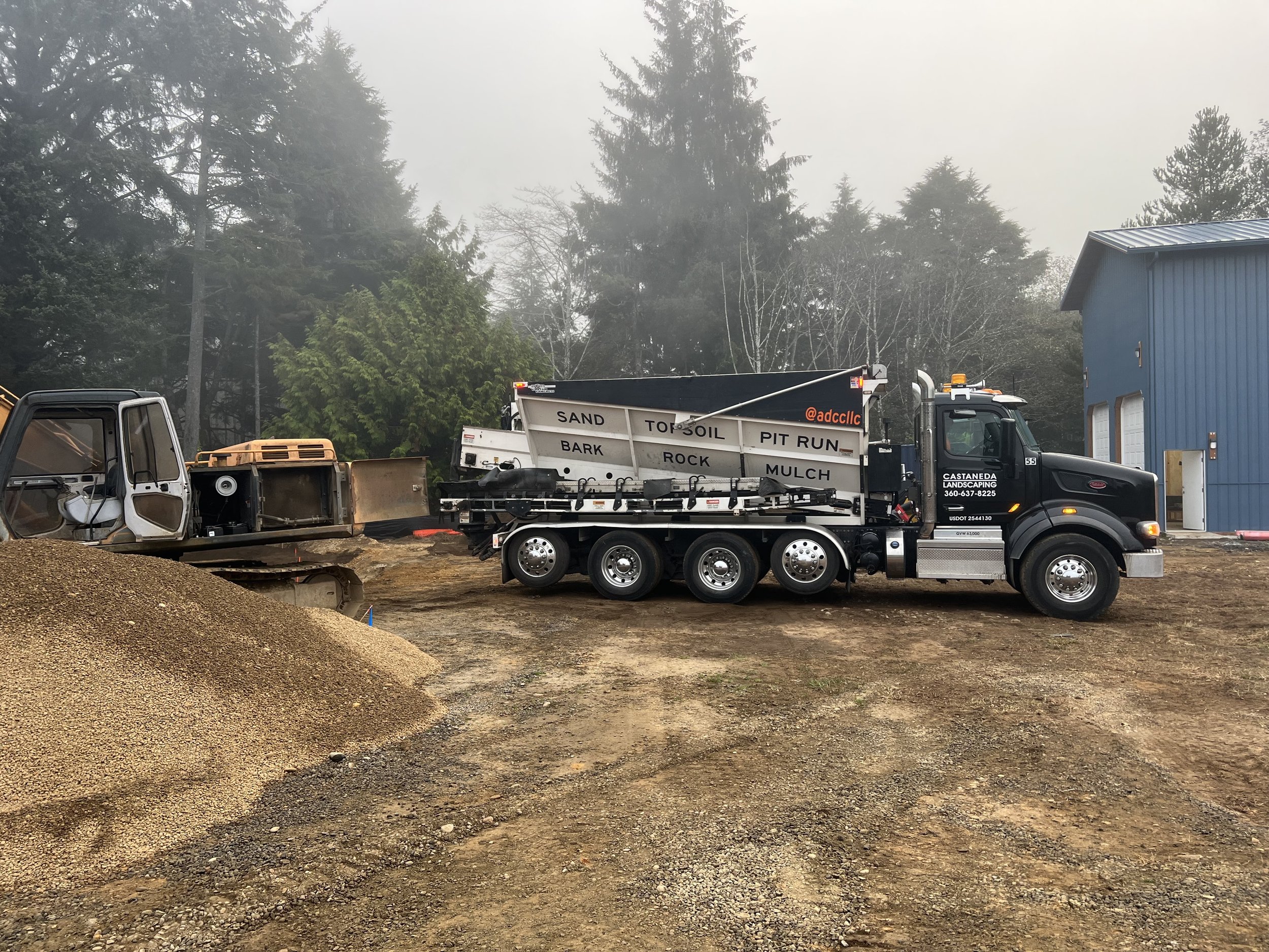 Construction site with a black dump truck loaded with gravel, a black truck labeled 'Casteneda Landscaping,' and various construction materials, set against a backdrop of green trees and a blue building.