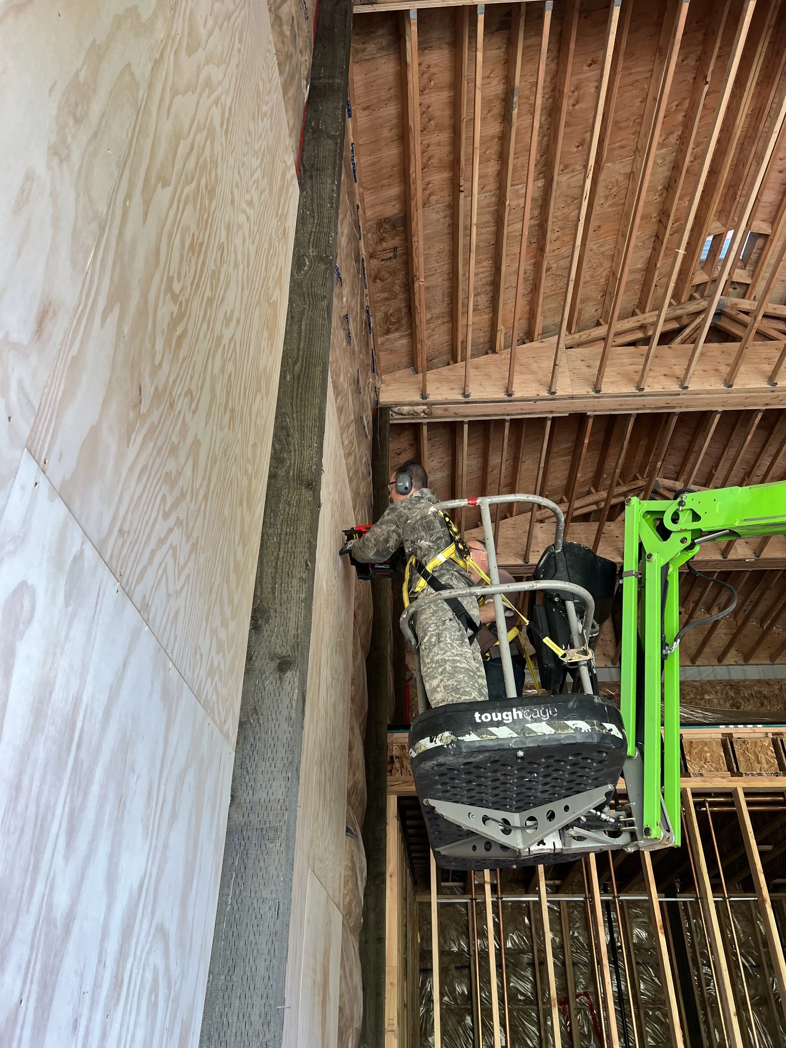 A worker in camouflage clothing is operating a green scissor lift inside a wooden structure under construction. The structure has exposed wooden framing and a plywood floor, with some plastic sheeting visible in the background.