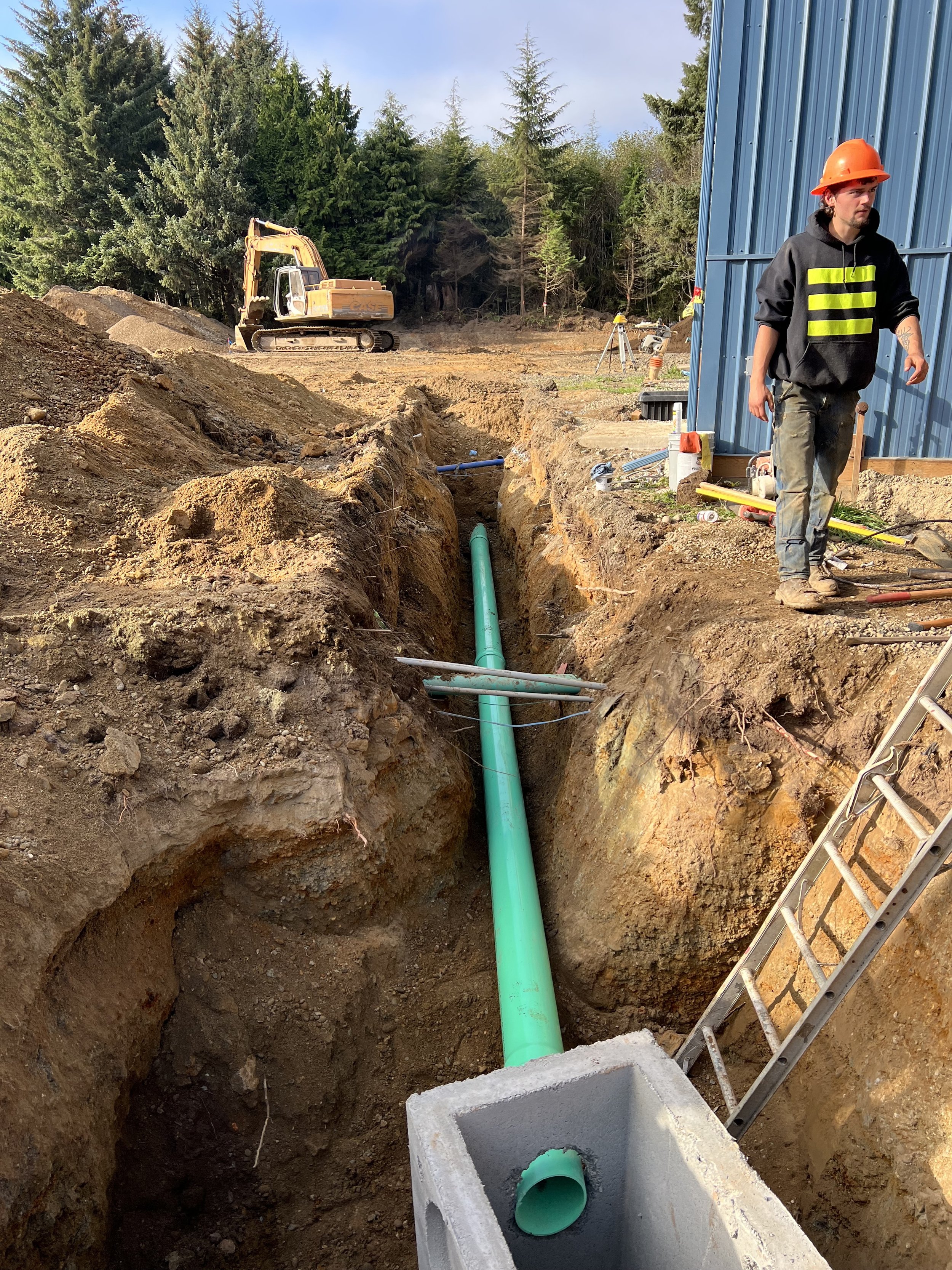 Construction site with an underground pipe installation, a worker wearing a hard hat and work clothes, a blue metal building wall, a yellow excavator in the background, trees, a ladder, and construction tools and materials.