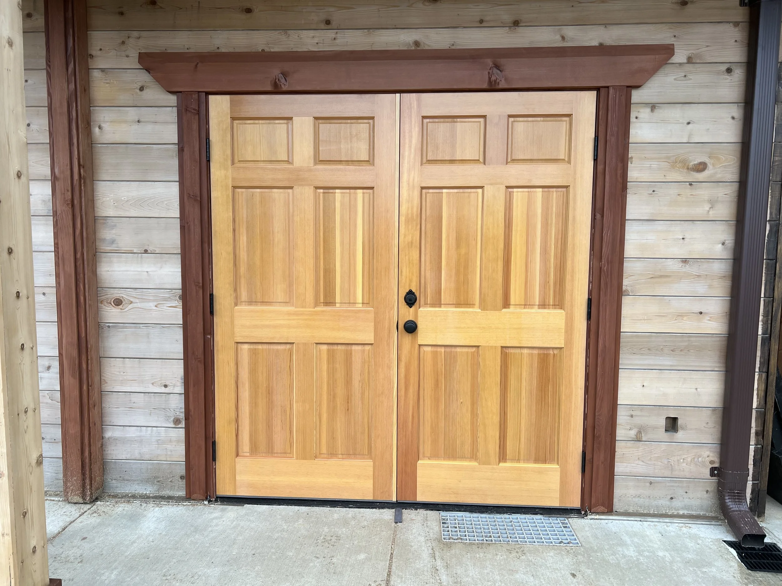 A double wooden garage door with a natural finish, surrounded by a wood-framed structure, set in a concrete driveway with a metal drain grate at the bottom.