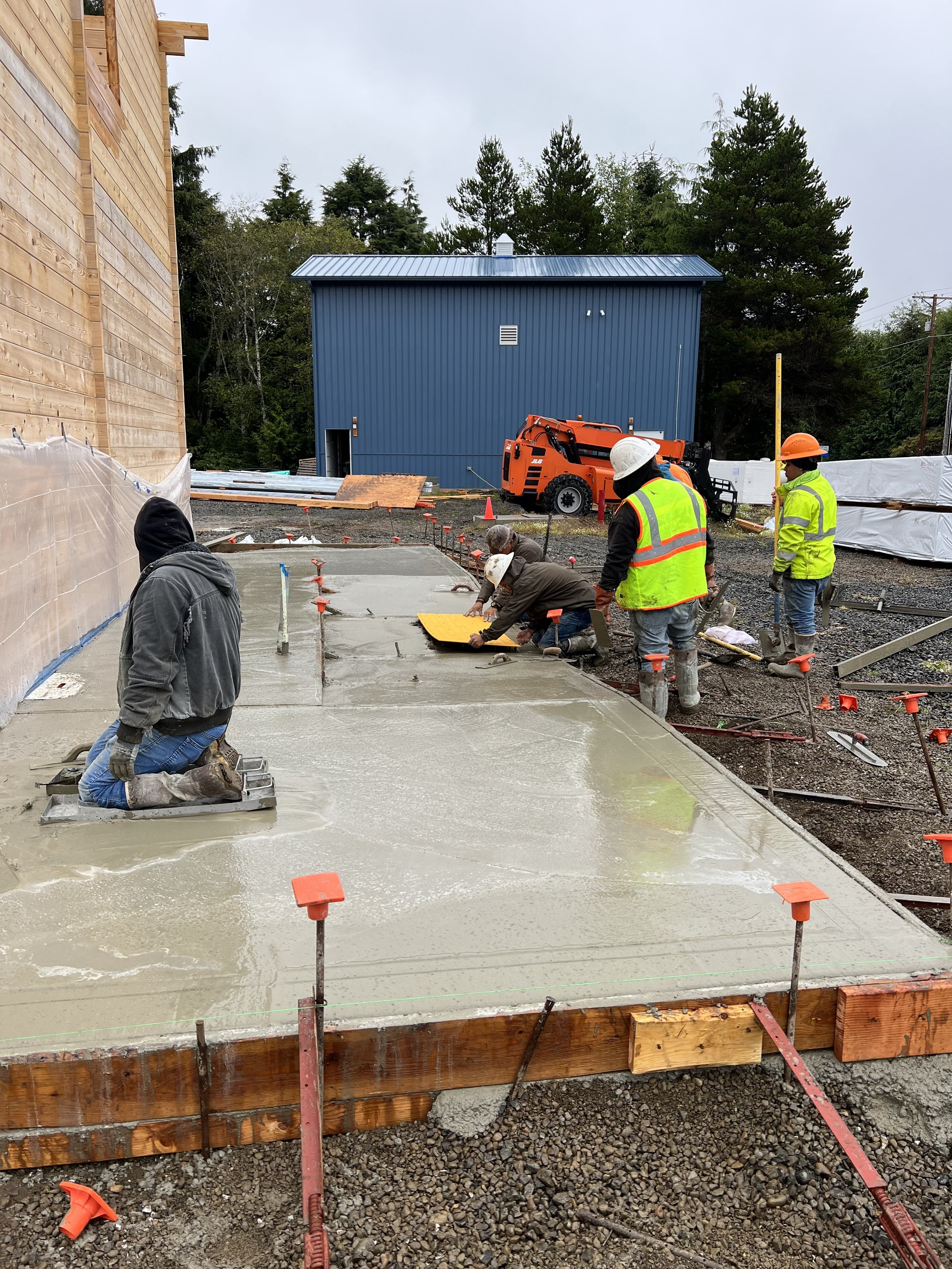 Workers pouring and leveling concrete on a construction site for a building foundation, with some workers kneeling and others standing, tools, and construction equipment nearby.