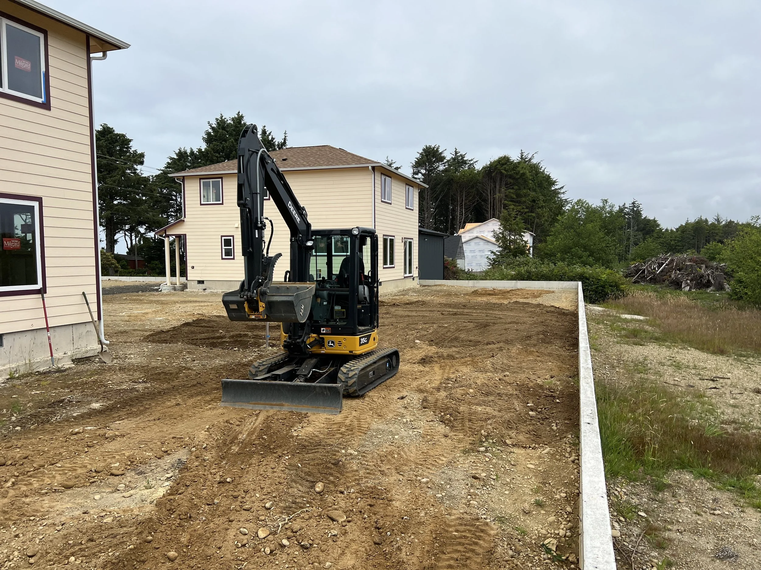 Construction site with a small excavator on soil ground, in front of residential houses, and trees and cloudy sky in the background.