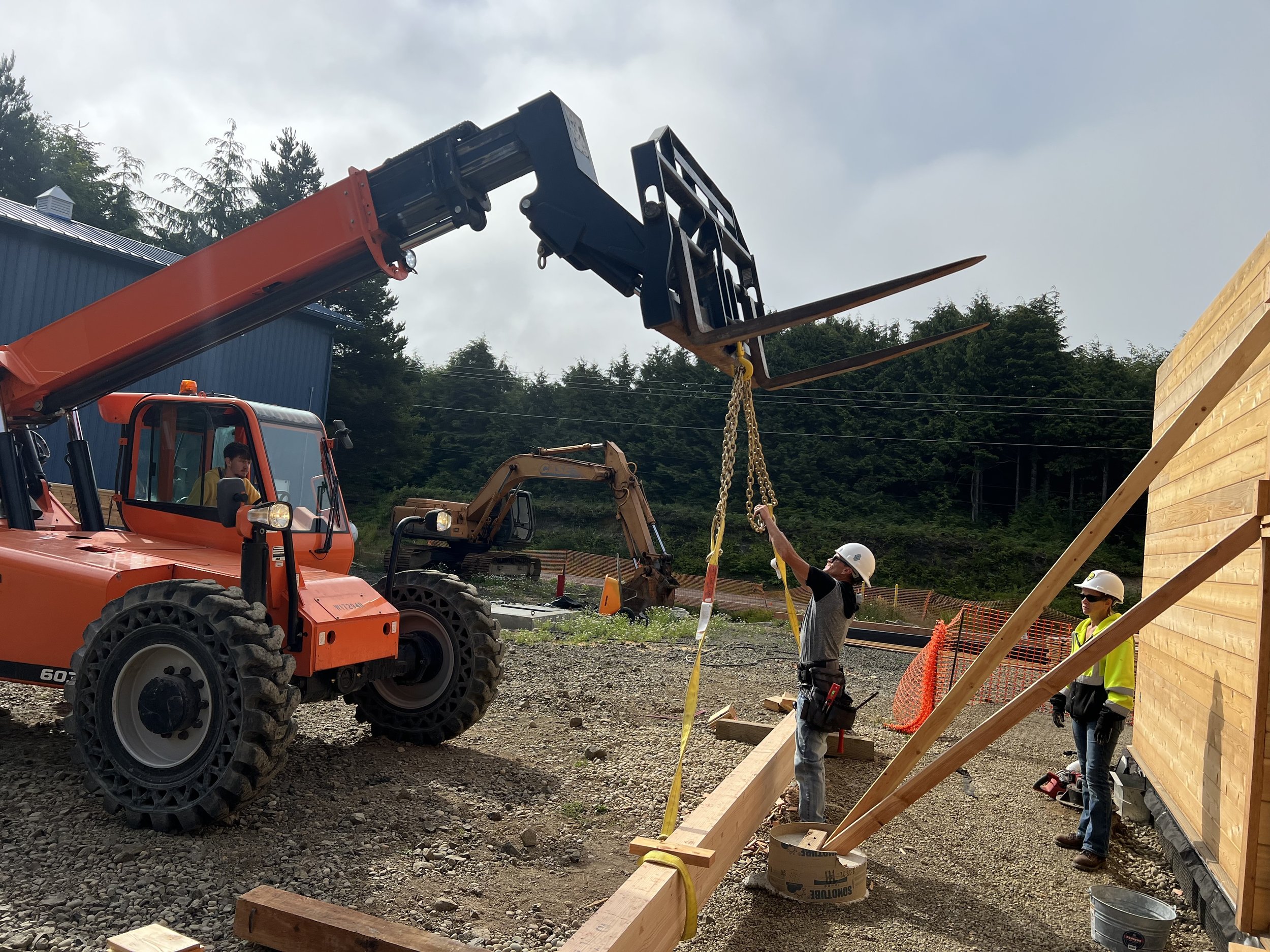 Construction workers and machinery lifting a wooden structure into place on a building site, with trees and cloudy sky in the background.