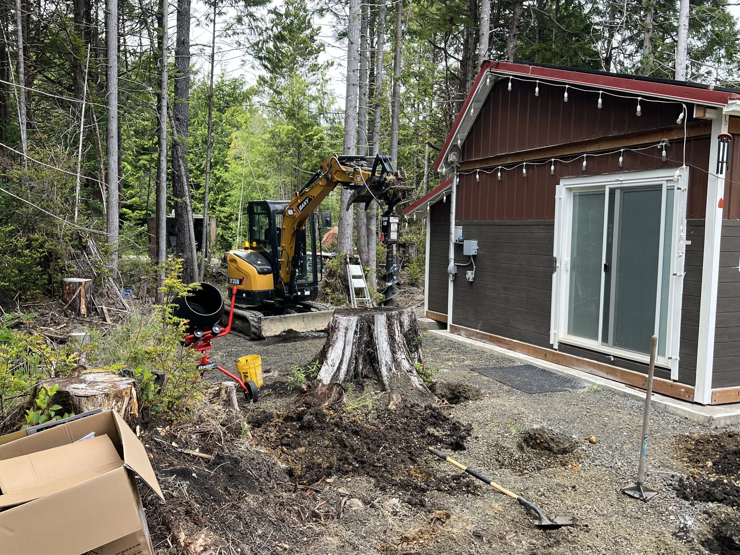 A backyard scene with a small excavator working near a house with brown siding and white trim, surrounded by trees and construction materials.