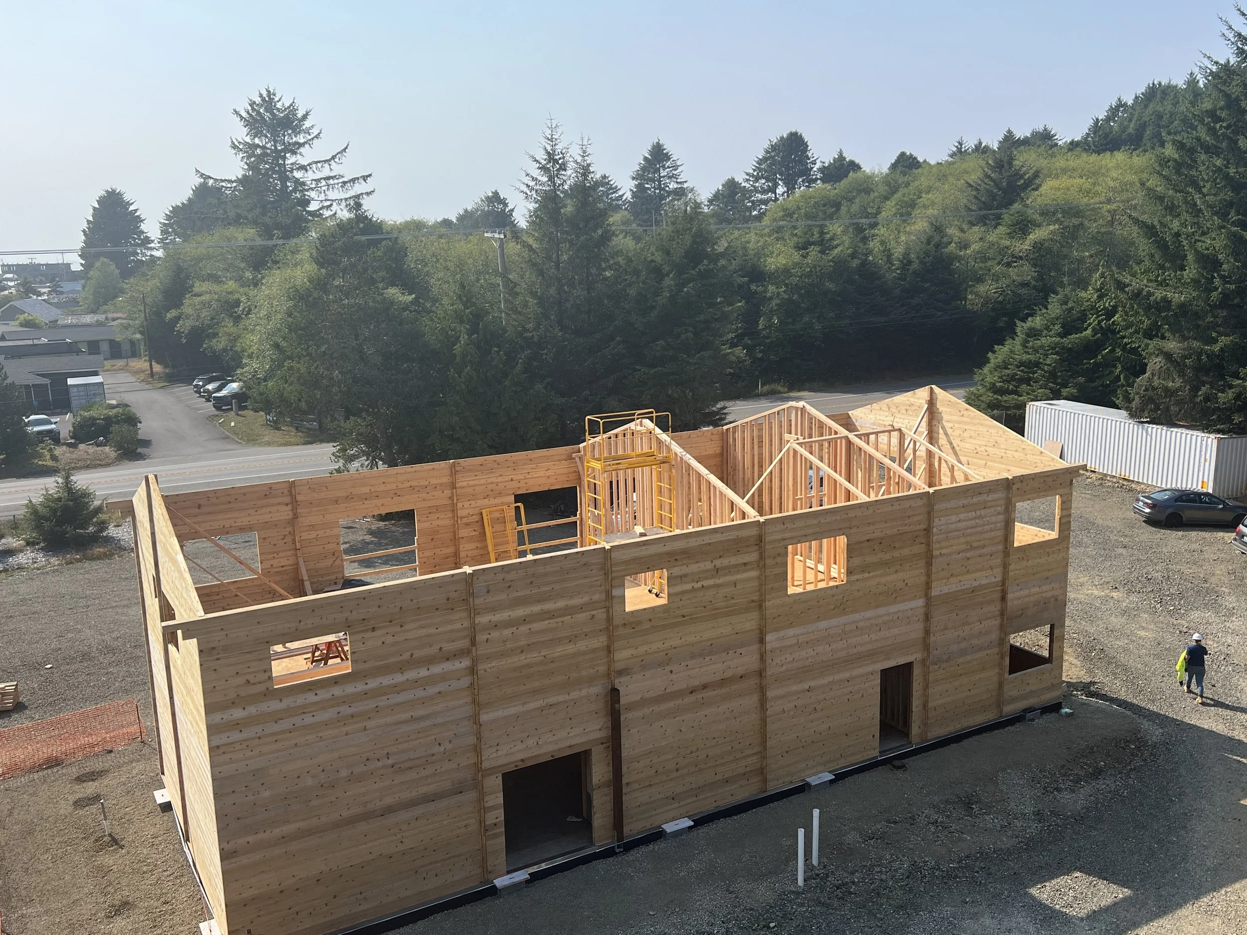 Construction site of a multi-story building with unfinished wooden walls and black foundation, surrounded by gravel and a worker in a yellow vest and helmet. In the background, there is a forested area with tall trees and a clear sky.