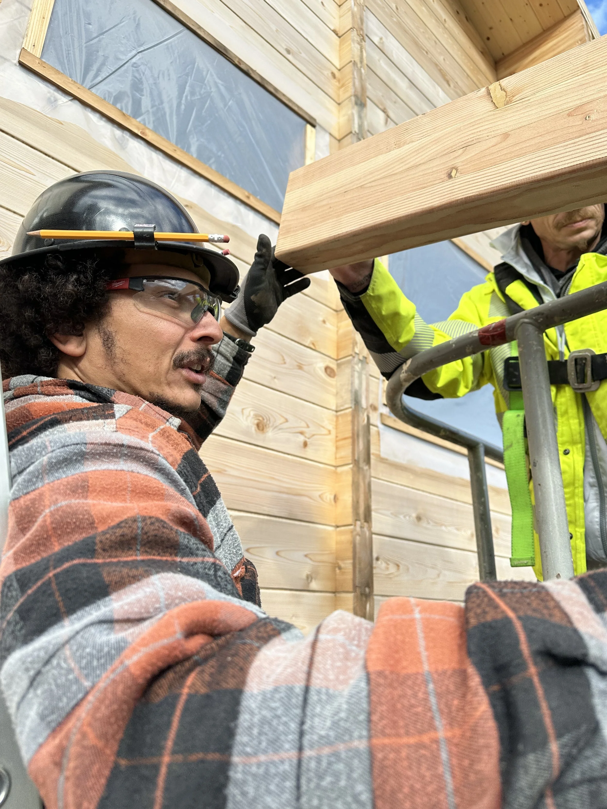 Two construction workers installing wooden wall panels on a building exterior during daytime. One worker with curly hair, beard, safety glasses, helmet, and plaid shirt is holding the panel, while the other in a neon yellow safety vest is assisting.