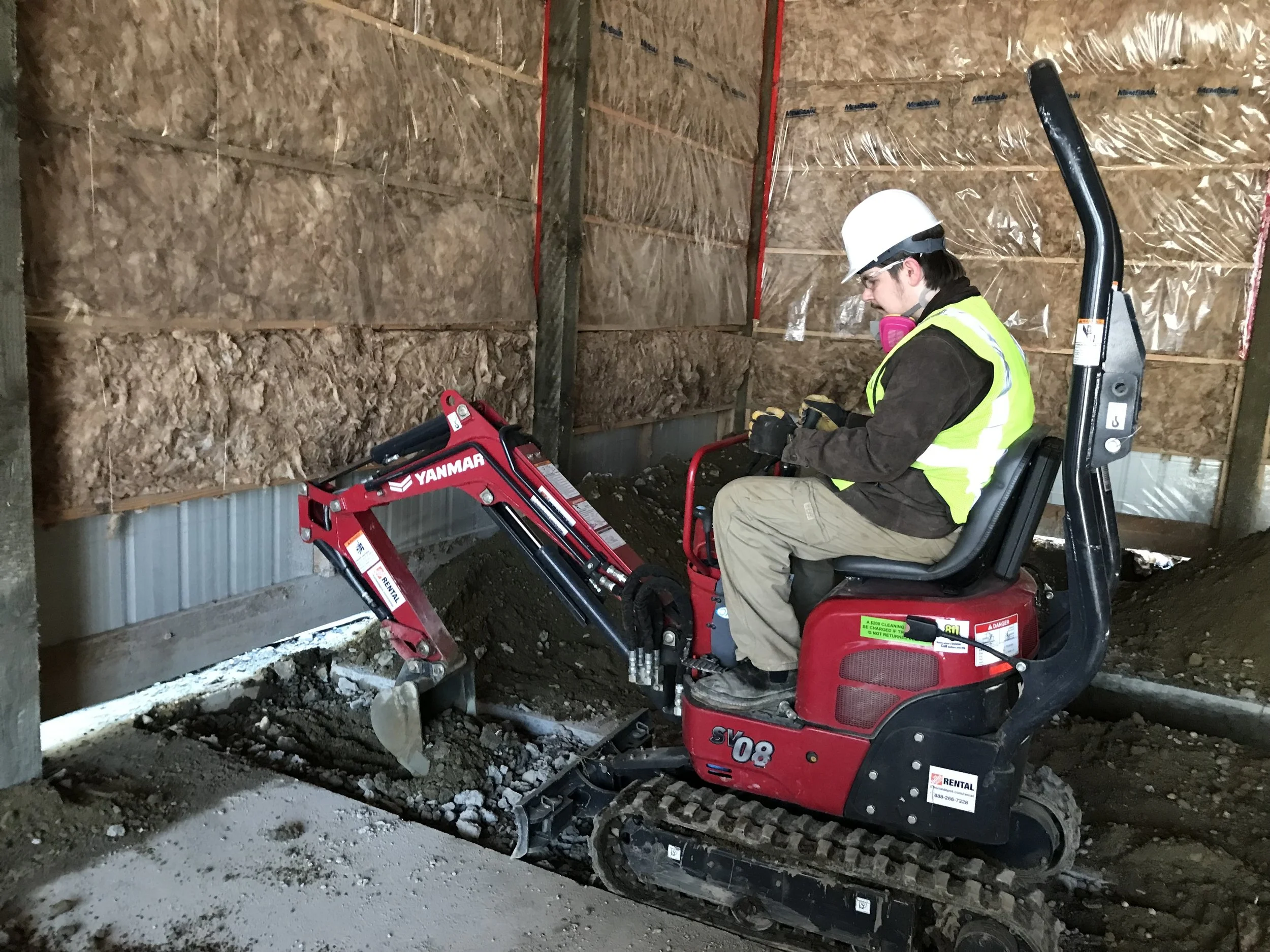 Construction worker operating a red mini excavator inside a building with unfinished walls covered in insulation and plastic sheeting.