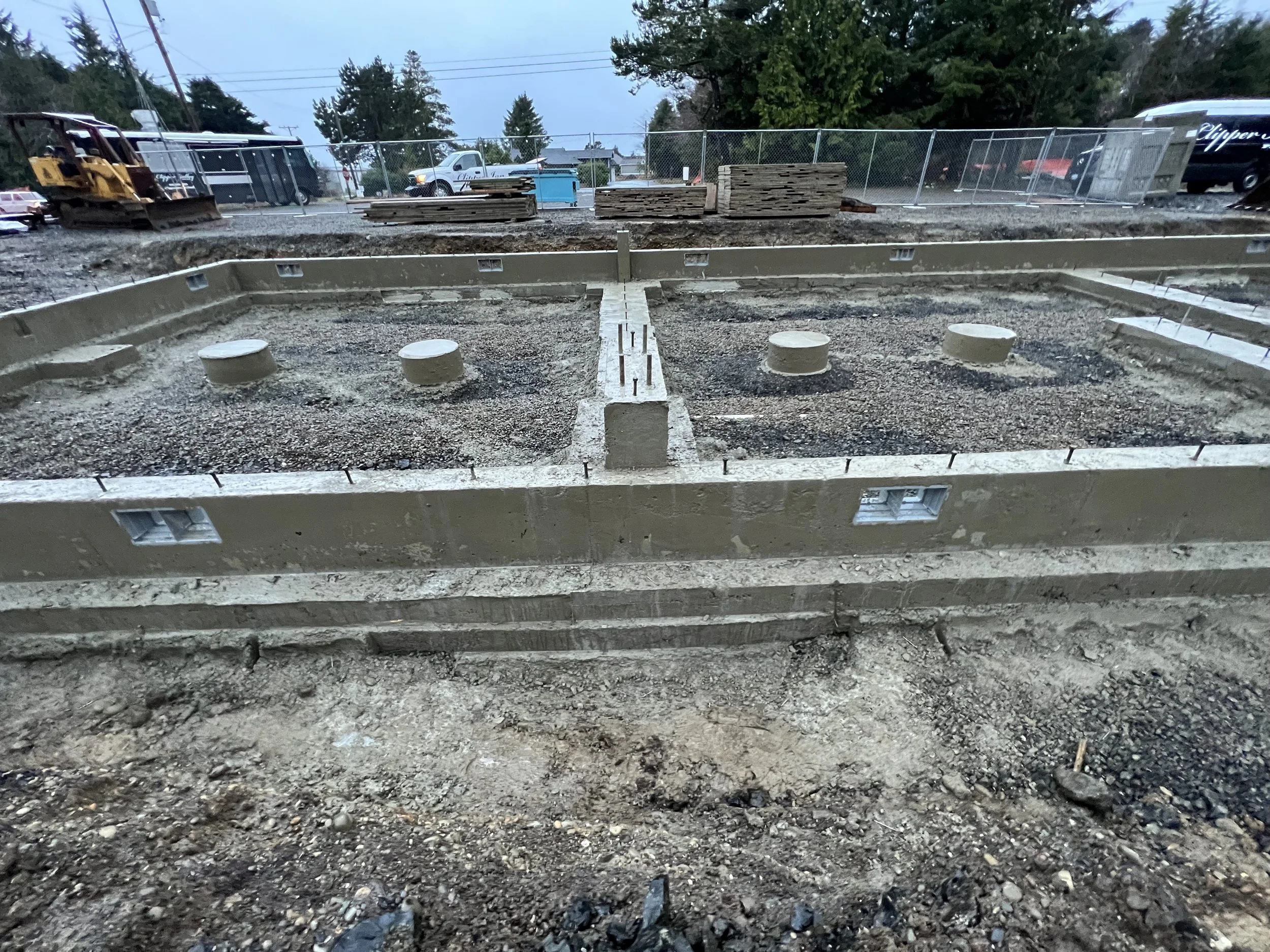 Construction site with foundation framing, concrete blocks, and construction equipment. Background with vehicles and trees during dusk.