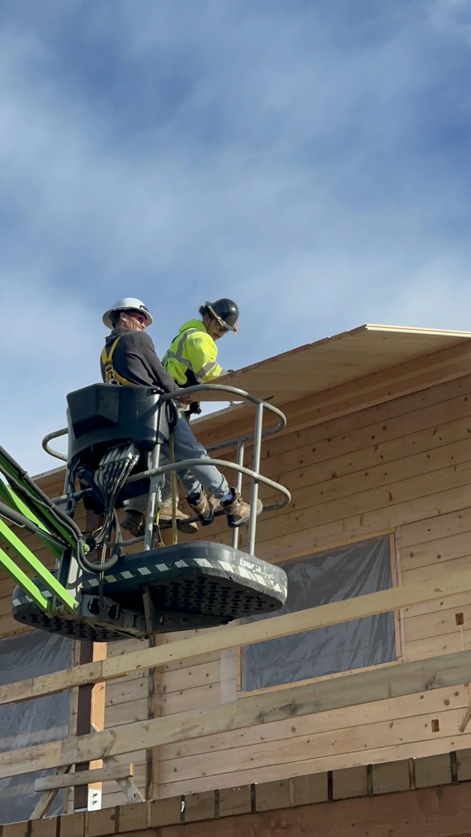 Two construction workers are on a lift working on the roof of a house. One worker is wearing a white helmet and the other is wearing a black helmet, both are in safety gear. The house is in the process of being built, with wooden siding and construct
