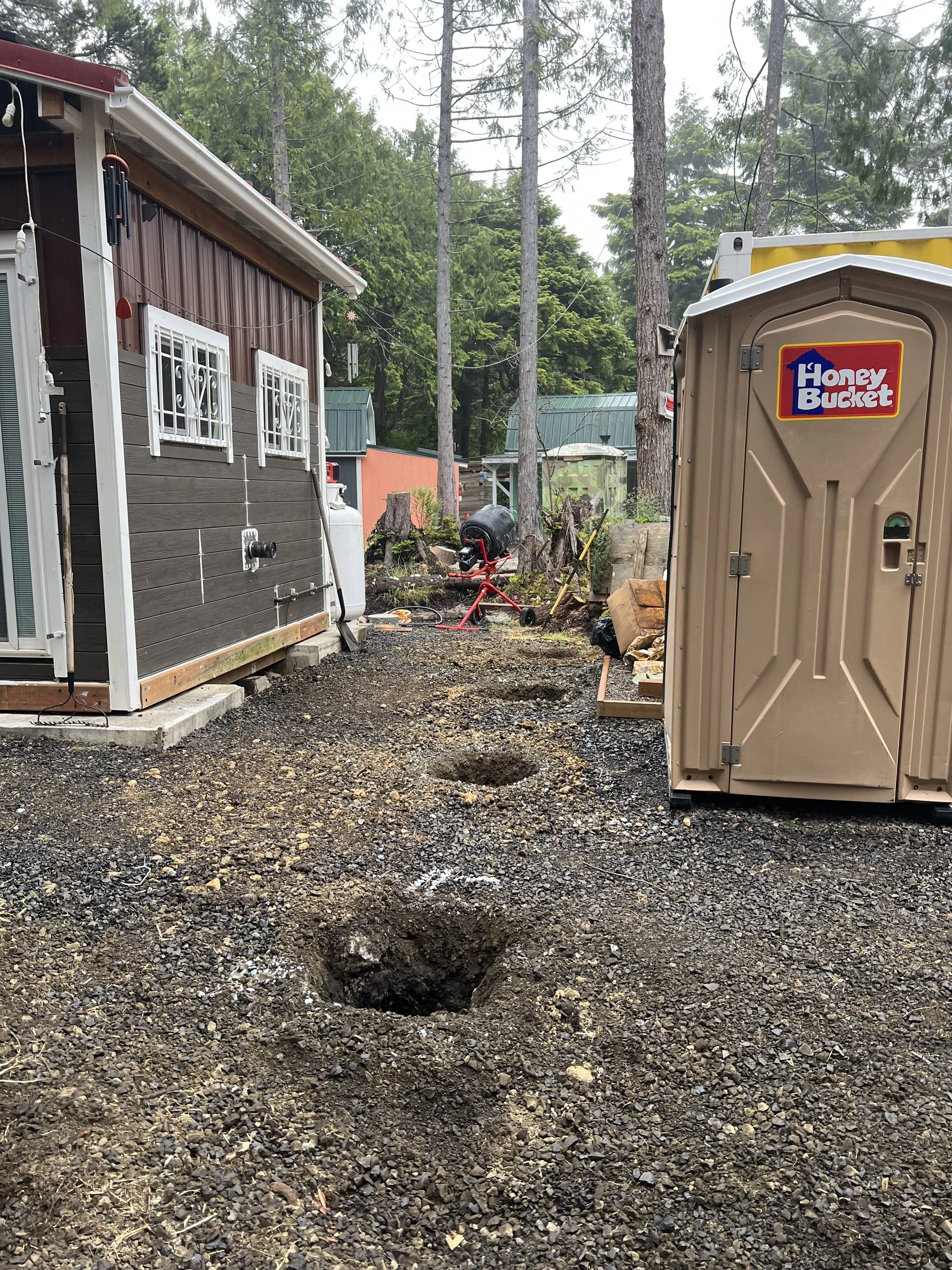 A dirt yard with a large hole in the ground, a storage shed on the right, a portable toilet labeled 'Honey Bucket' on the right, and trees in the background.