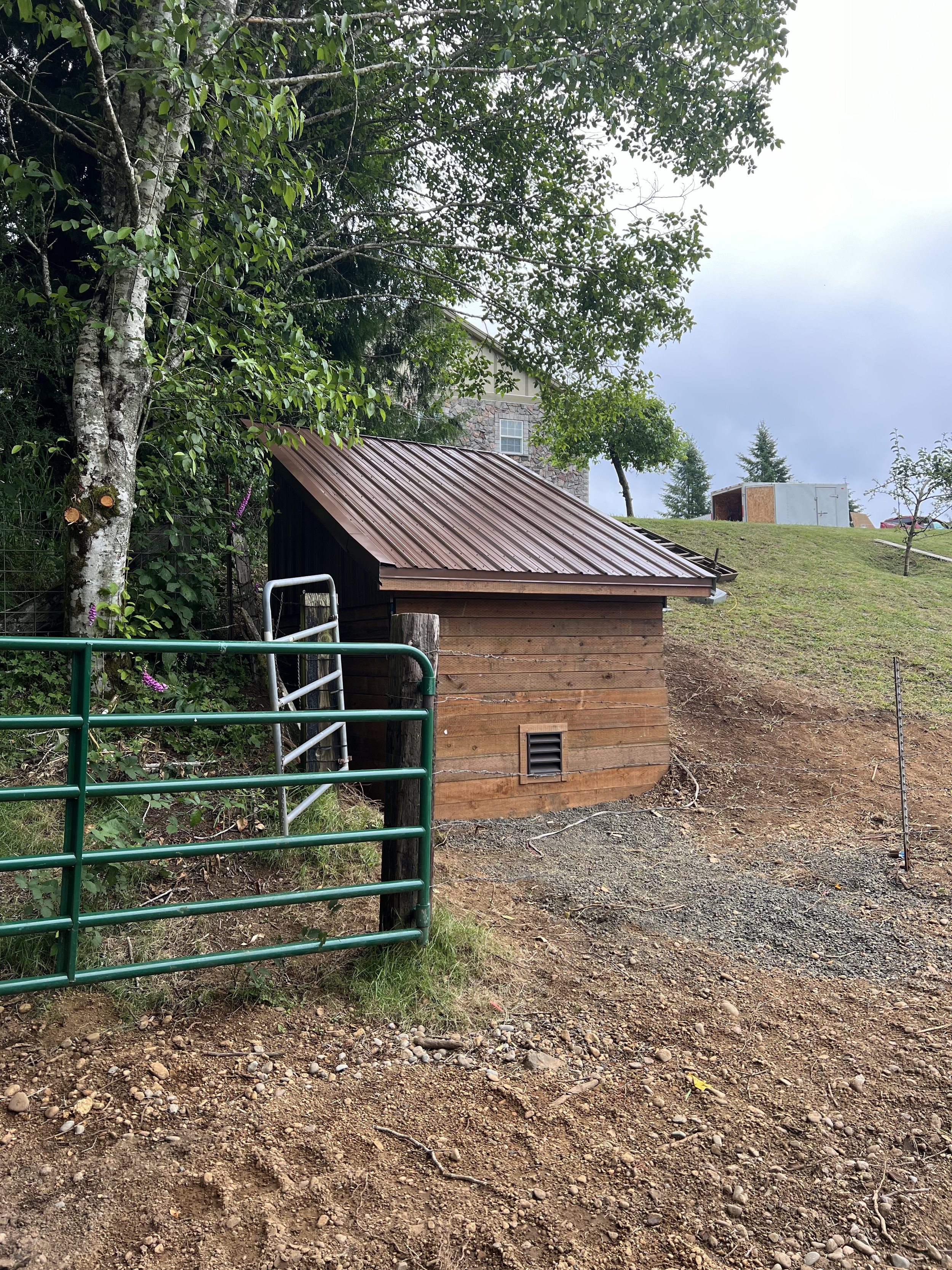 A small wooden shed with a brown metal roof, situated on a grassy hill, partially obscured by trees and greenery, with a gravel path in front and a green metal gate.