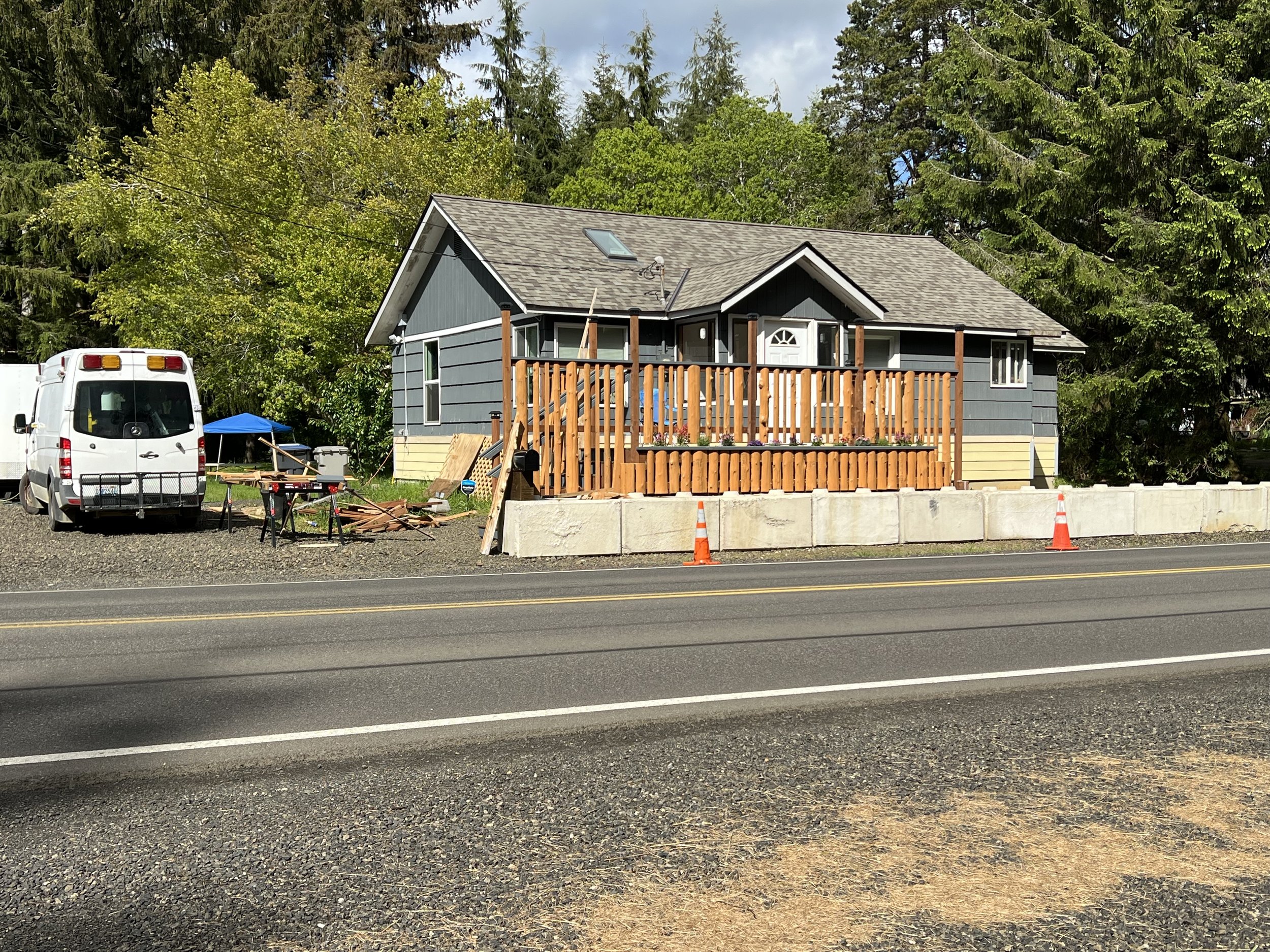 A blue house with a wooden fence under construction, parked white van, and construction tools and materials, with a backdrop of green trees and a partly cloudy sky.
