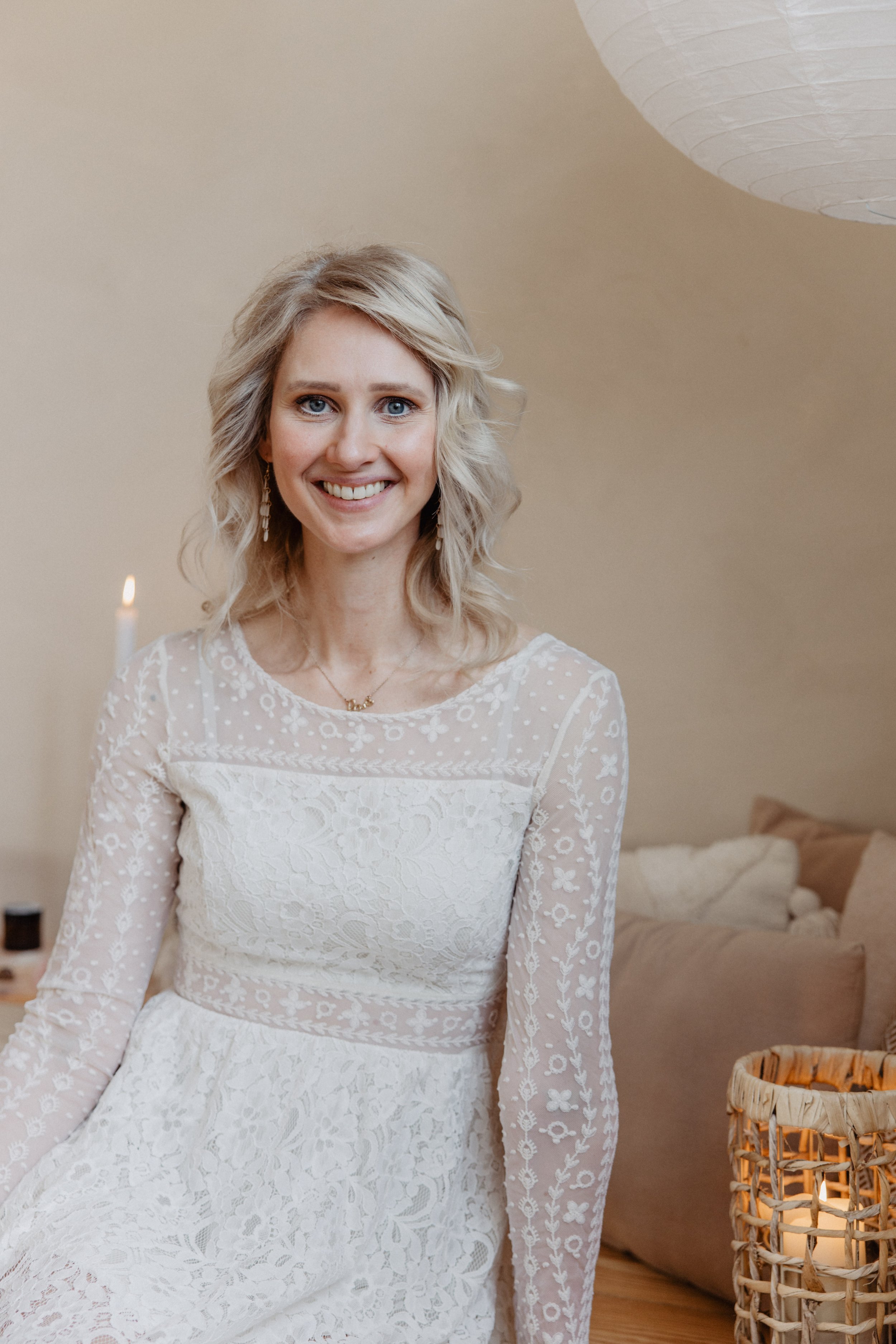 Ein lächelndes blondes Mädchen in einem weißen Spitzenkleid, im Hintergrund eine beige Wand, ein Papierlampenschirm und eine Stehlampe, auf einem Couchtisch befinden sich Kerzen und Kissen.