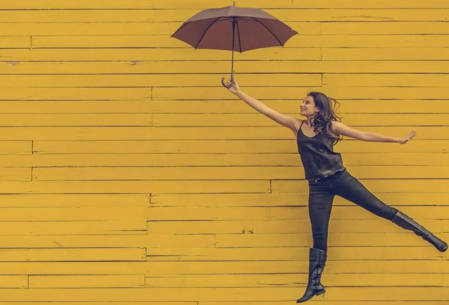 Woman in black outfit holding an umbrella, jumping in front of a yellow wooden wall.