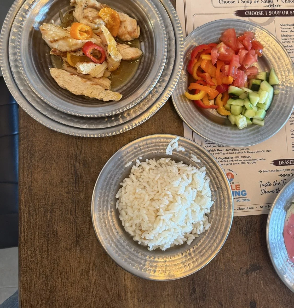Hammered metal plates of grilled chicken, a tomato and cucumber salad and rice on a wooden table at Clay Pot in Naples