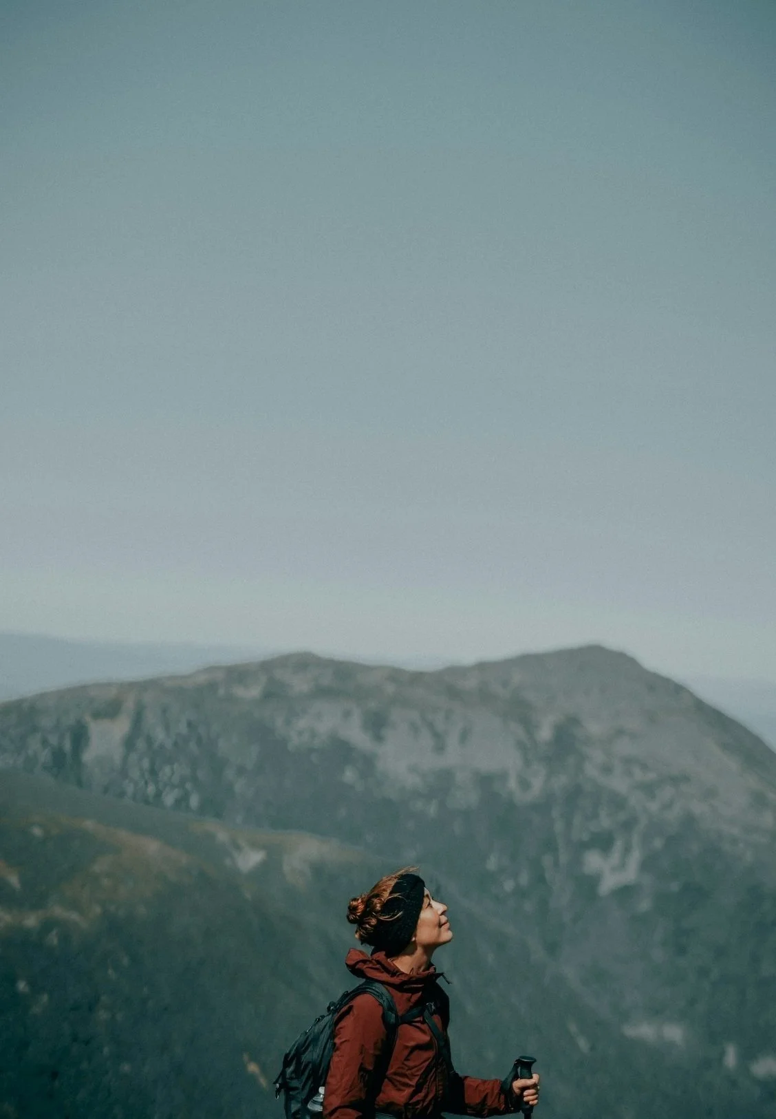 A woman hiking in the mountains, wearing a red jacket and black headband, carrying a backpack and holding a trekking pole, with mountain peaks in the background.