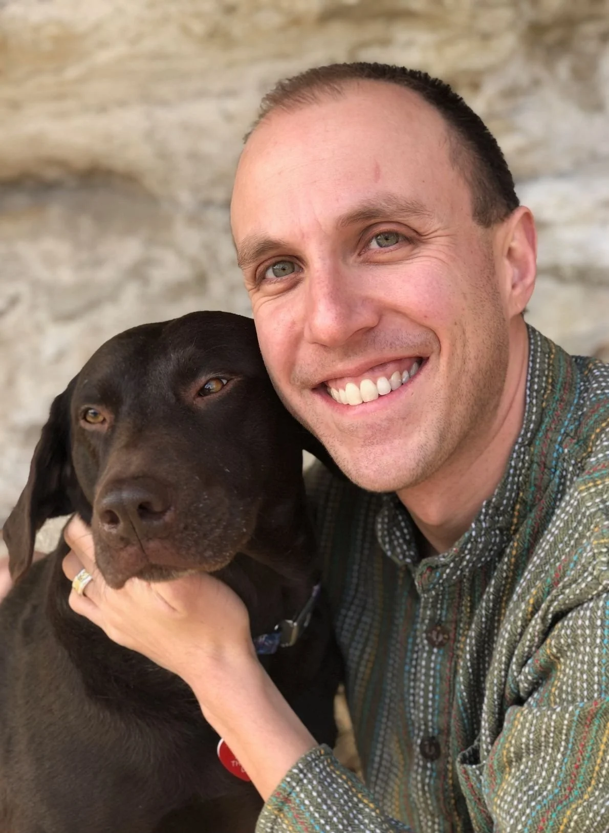 Matt Angleman, therapist in Grand Junction, CO, smiling and holding a black dog close to his face, both against a stone wall background.