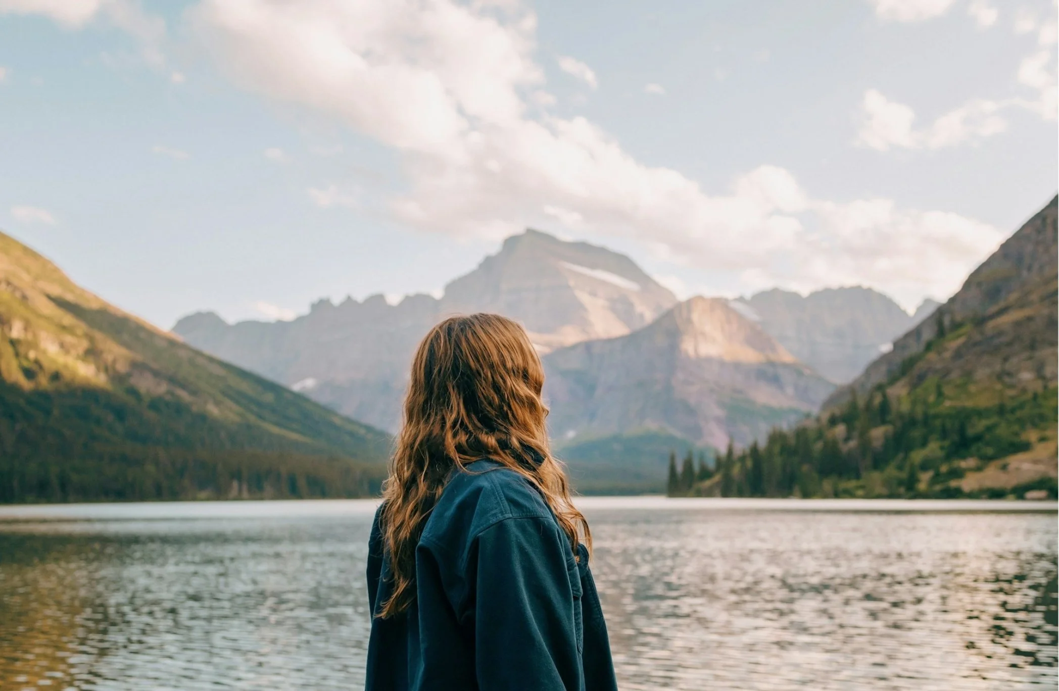 A woman with long brown hair wearing a blue jacket standing in front of a lake with mountains in the background.