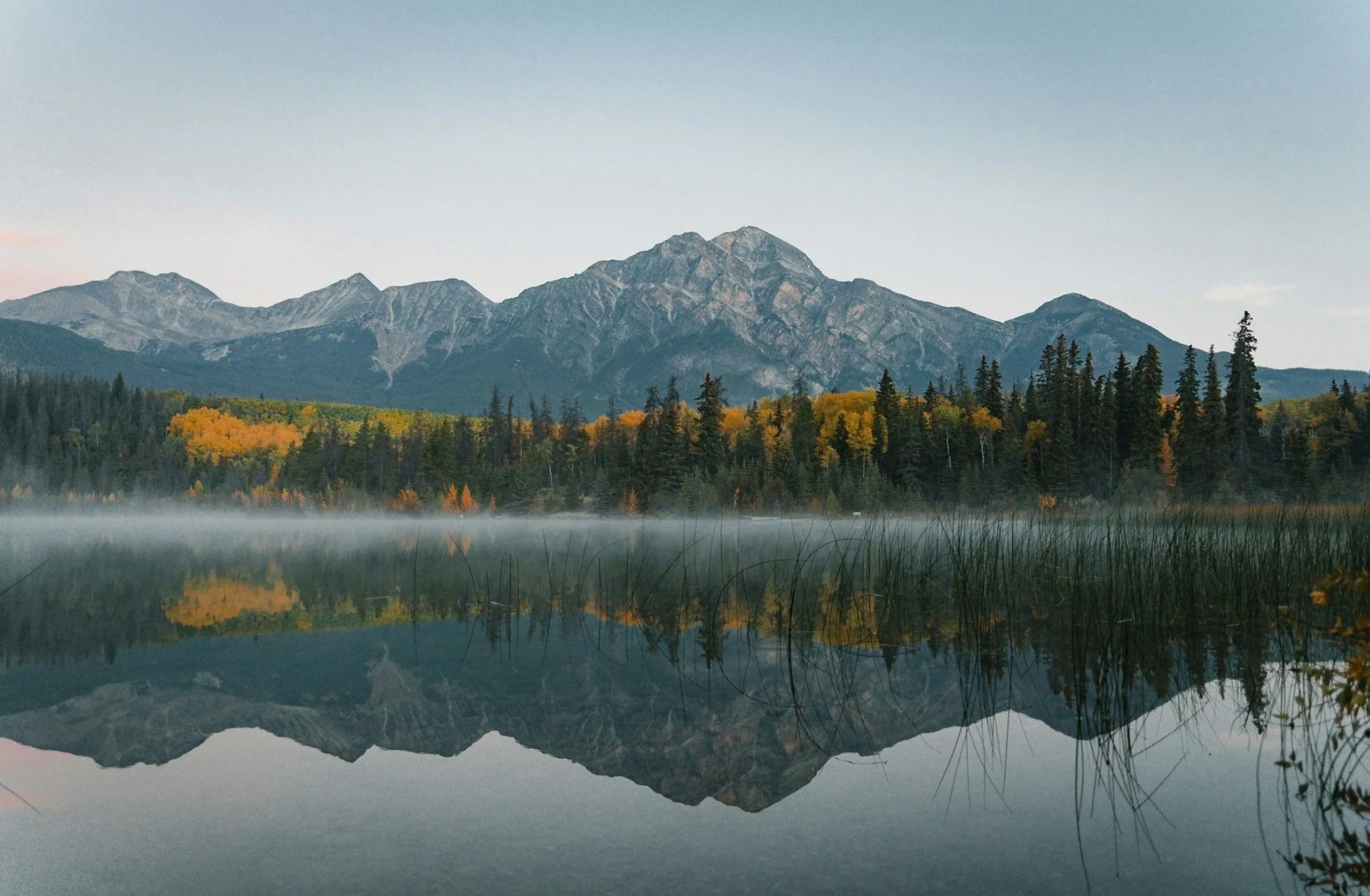 A mountain range reflected in a calm lake surrounded by trees with autumn foliage.