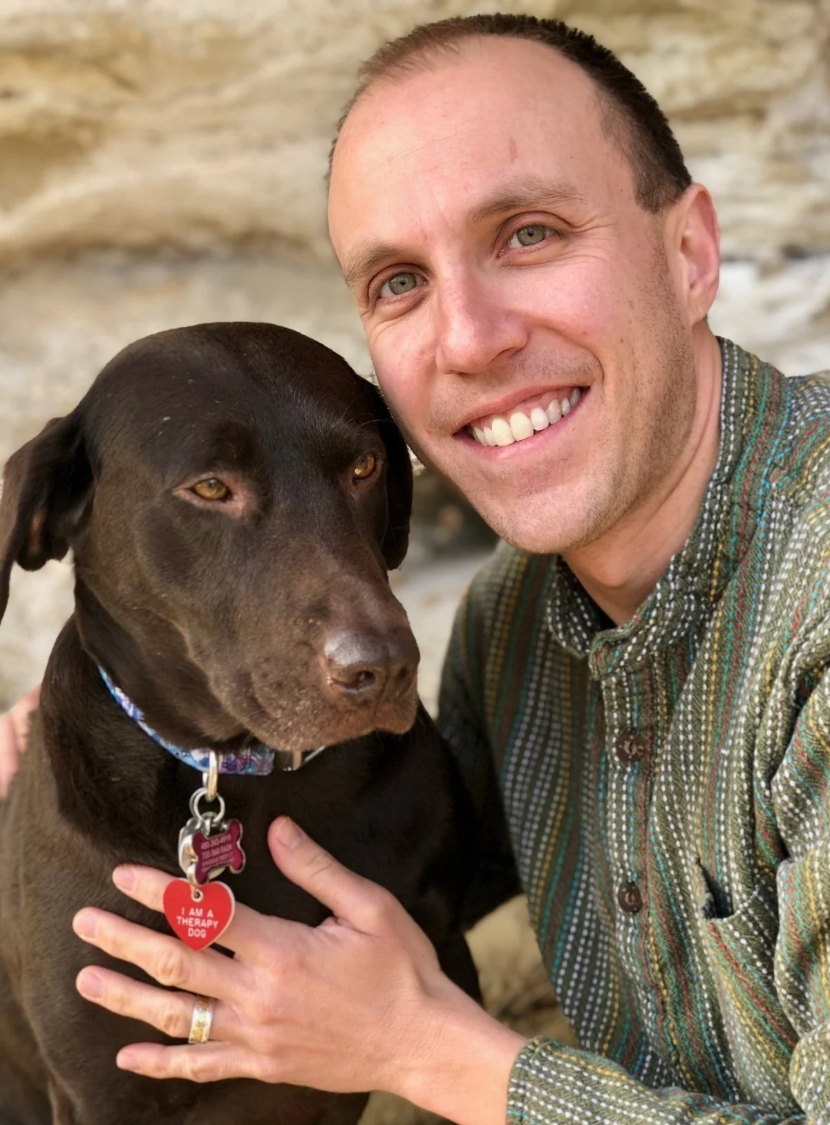 Matt Angleman, therapist in Grand Junction, CO, smiling with light skin, short hair, and a patterned shirt taking a close-up selfie with a brown therapy dog that has a blue collar and tags. The background is rocky.