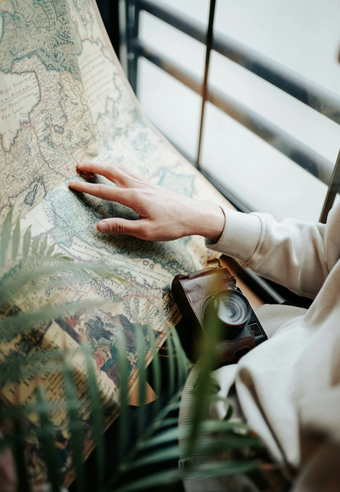 Person pointing at a historical map on a table, with a camera hanging from their wrist and some green plants in the foreground.