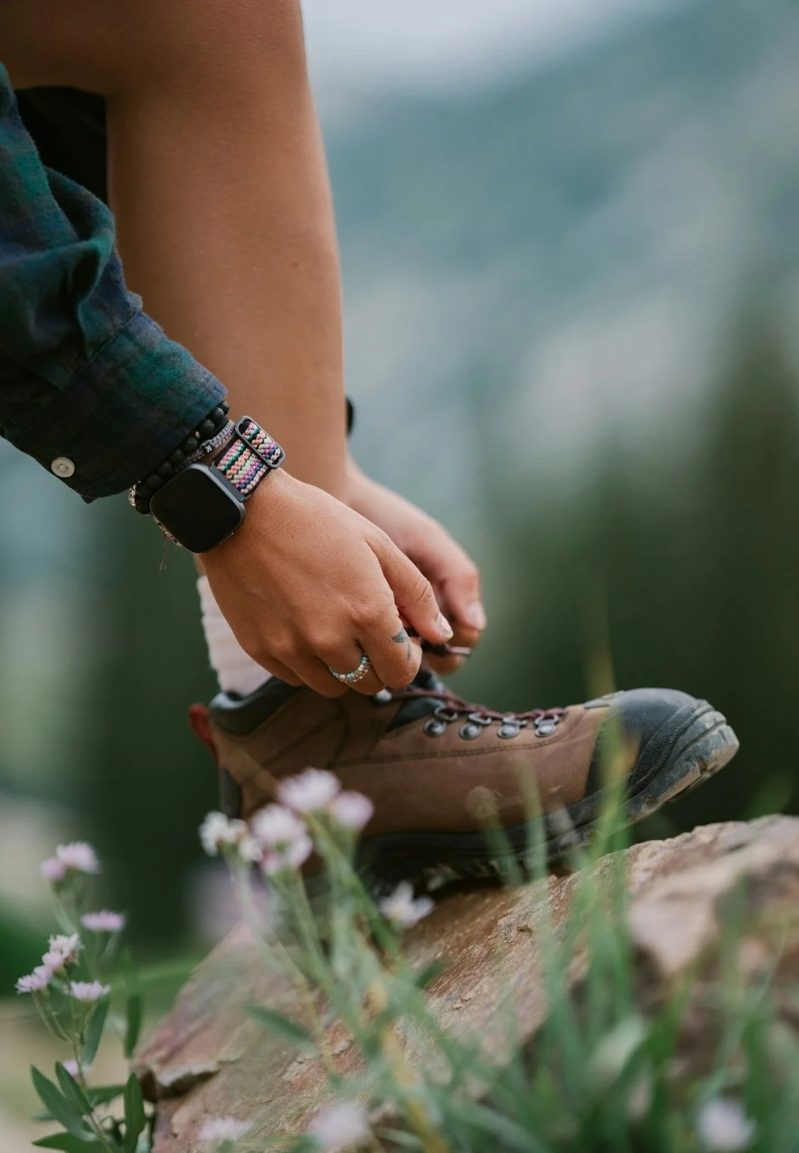 Person tying hiking boot outdoors surrounded by green plants and small pink flowers.