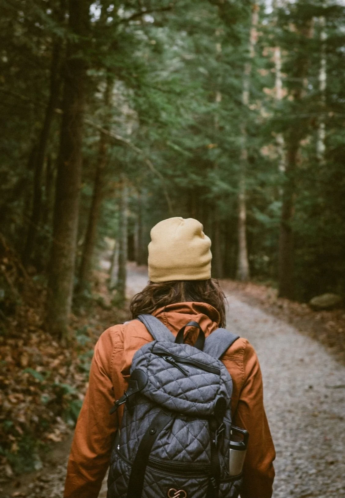A person with brown hair wearing a beige beanie, orange jacket, and carrying a gray backpack walking on a trail in a forest with tall green trees.