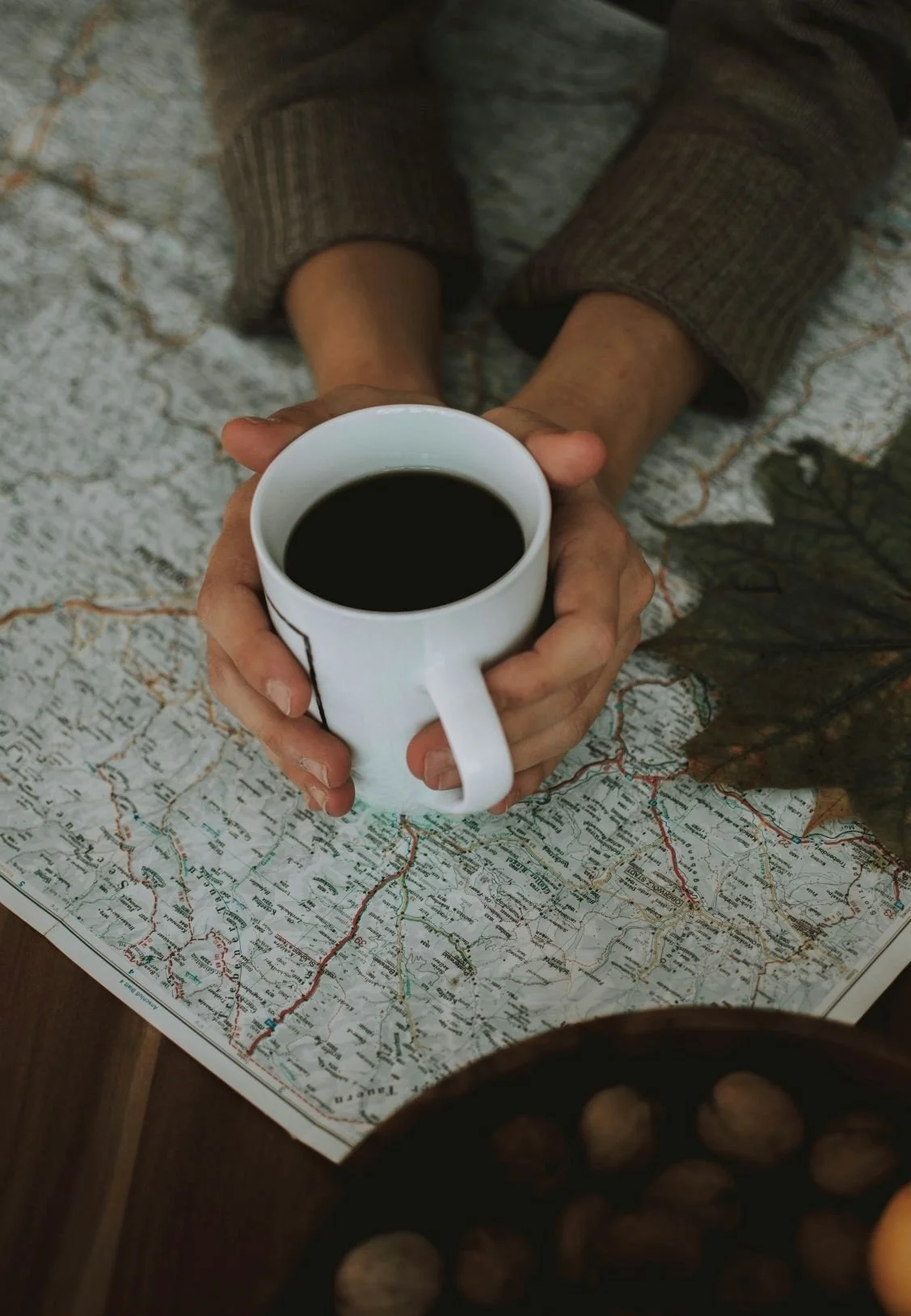 Person holding a white mug filled with coffee over a map on a table, with a bowl of nuts in the foreground.