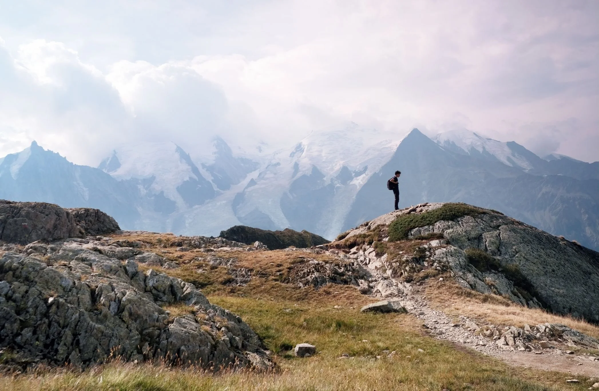 A person standing on rocky terrain in a mountainous area with snow-capped peaks and cloudy sky in the background.