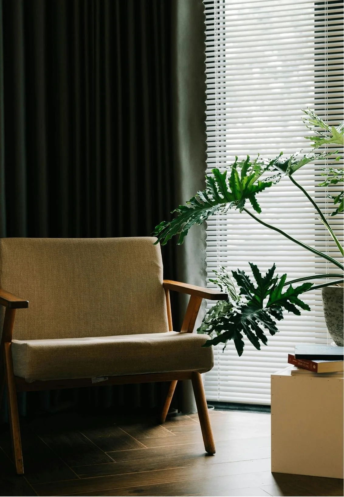 A living room corner with a beige armchair, green houseplants near a window with white blinds, and a small white side table with a stack of books.