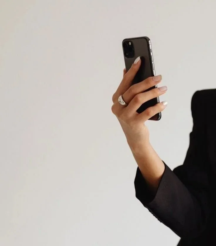 Person holding a smartphone with a black case, wearing a ring, in front of a plain light-colored background.