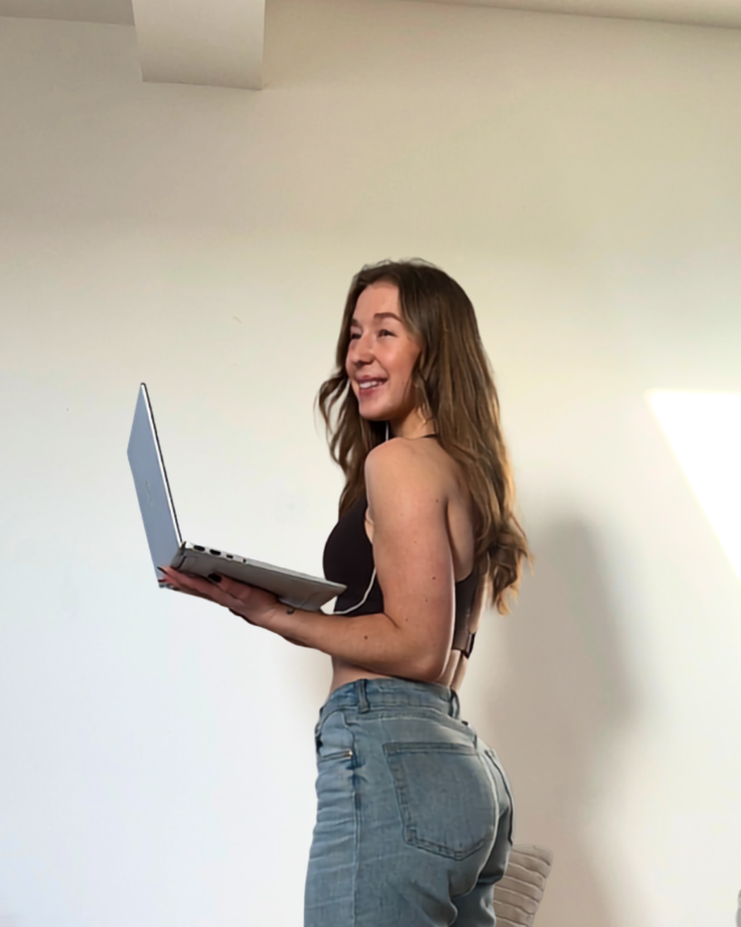 A woman with long brown hair, wearing a black sleeveless top and blue jeans, is smiling while holding a silver laptop in a bright room with plain white walls.