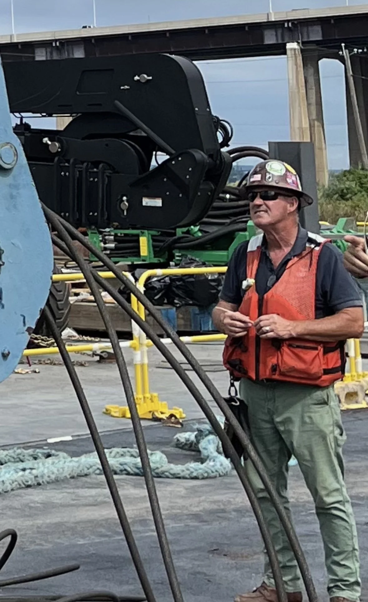 A construction worker wearing a helmet, sunglasses, a safety vest, and cargo pants stands outdoors near construction equipment, with a bridge in the background.