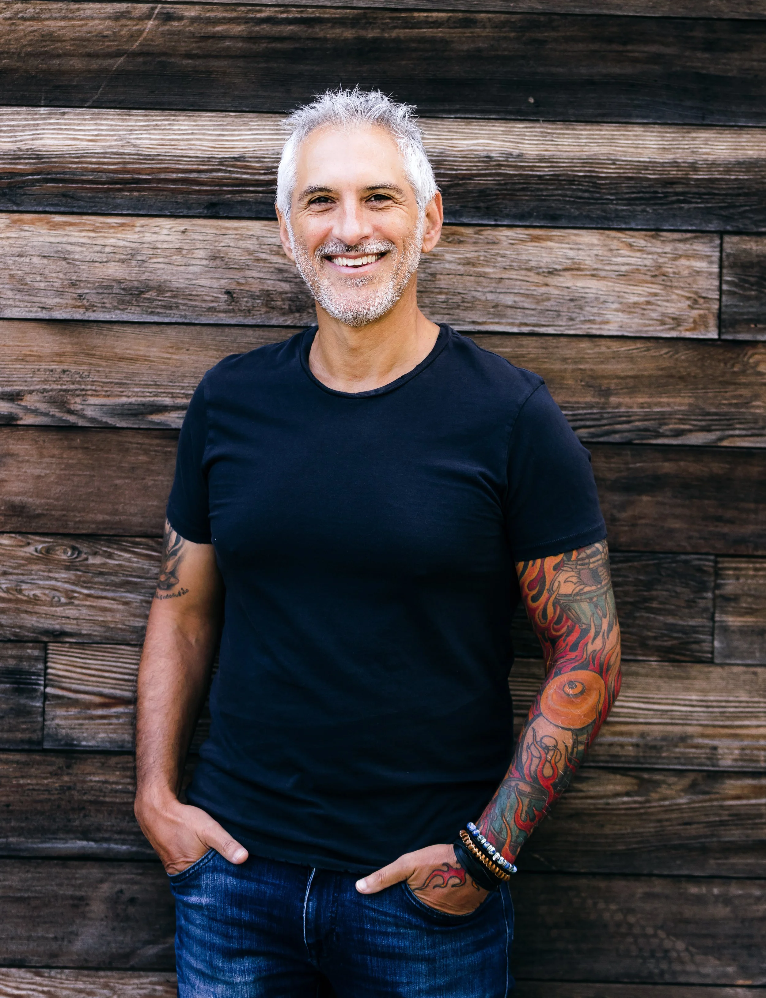 A middle-aged man with gray hair and a beard, smiling, wearing a black t-shirt and blue jeans, standing against a wooden fence background.