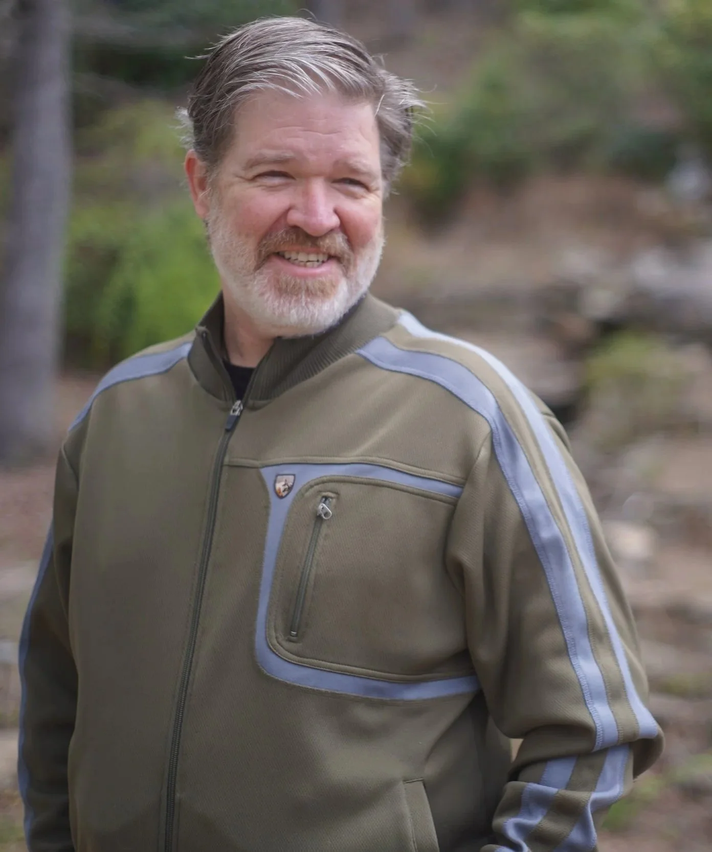 A smiling middle-aged man with gray hair and a beard outdoors in front of trees, wearing a khaki and gray jacket with patches.