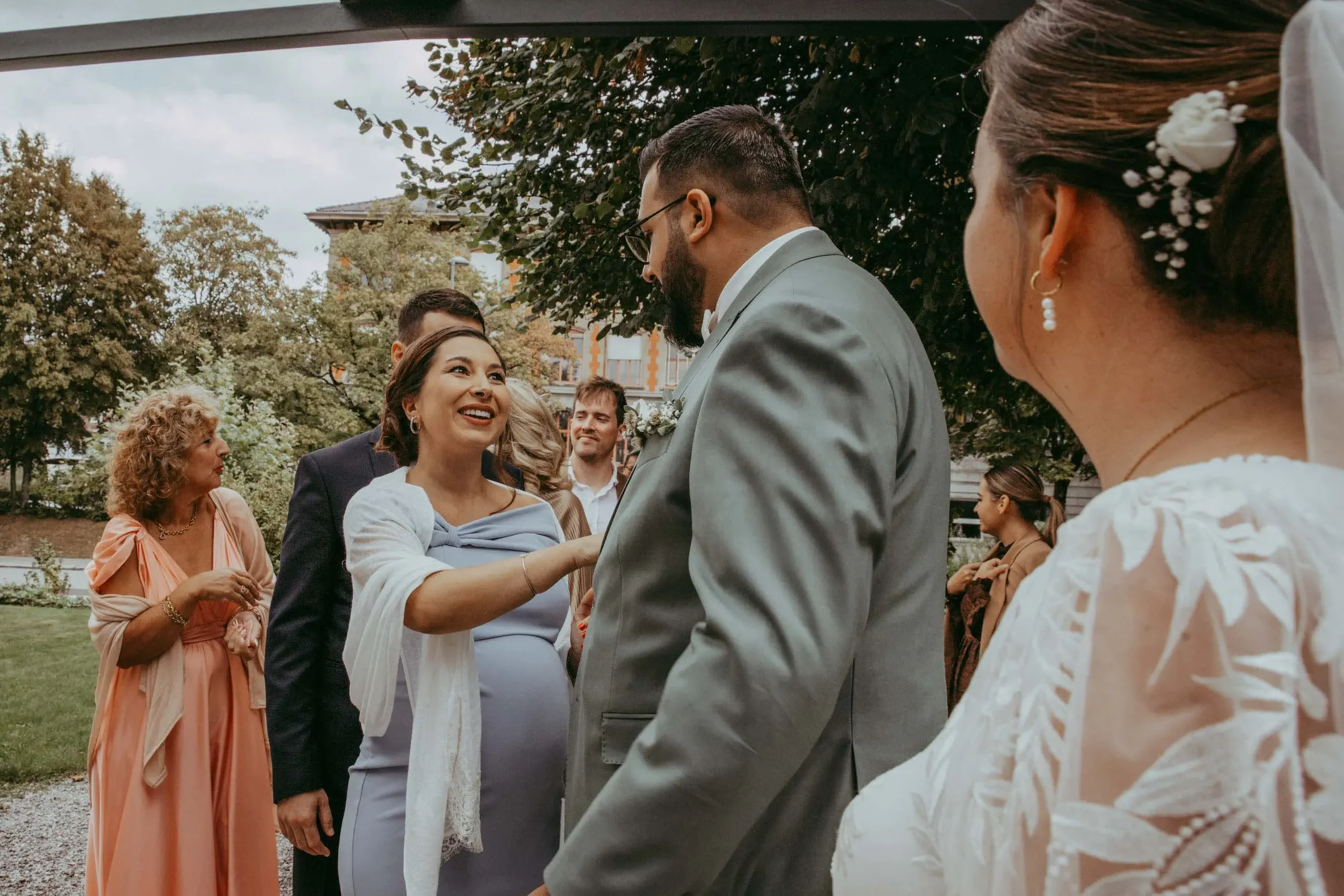 Hochzeit im Freien mit dem Brautpaar und Gästen, einige im Gespräch, im Hintergrund Bäume und ein Gebäude, bewölkter Himmel.