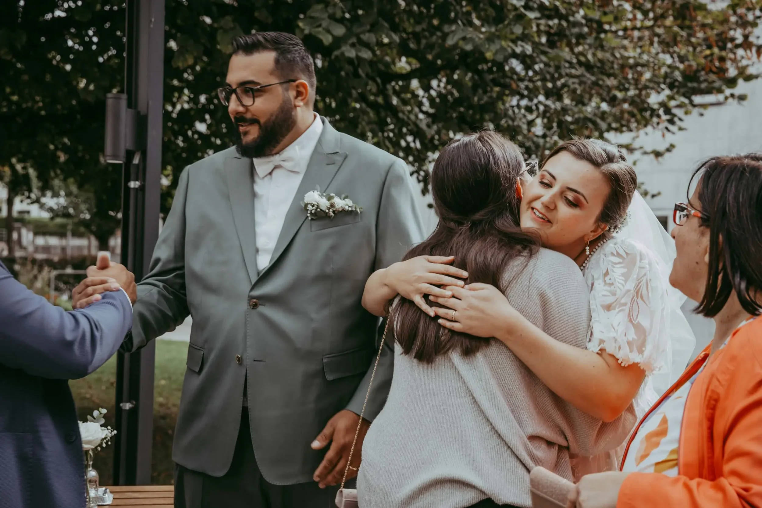 Menschen bei einer Hochzeit umarmen sich und begrüßen sich im Freien, bunte Blätter im Hintergrund.