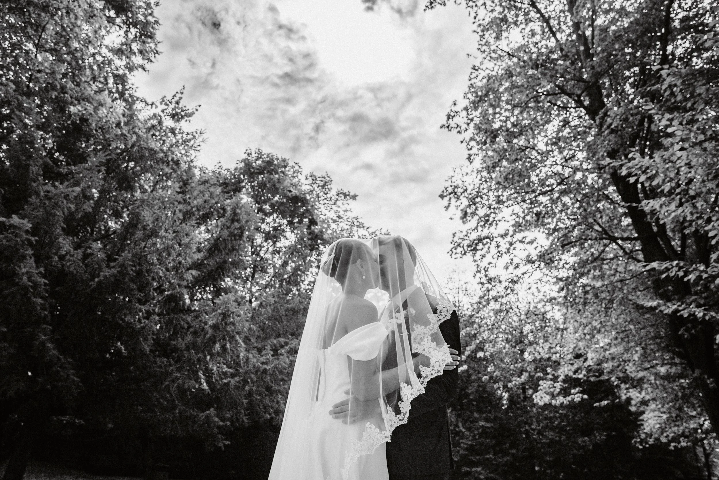 Ein Brautpaar in Hochzeitstracht steht sich im Freien gegenüber, umgeben von Bäumen, im schwarz-weißen Stil aufgenommen.