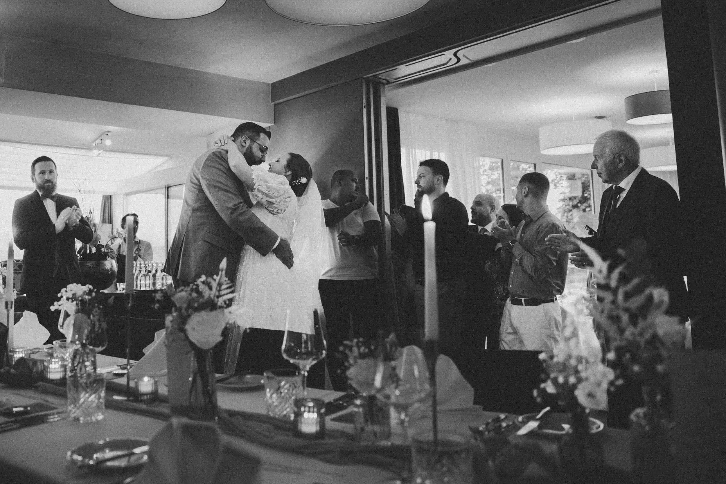 Ein Hochzeitsessen in einem modernen Raum mit mehreren Gästen, die die Hochzeit feiern, darunter das Brautpaar in der Mitte, das sich küsst. Der Raum ist hell beleuchtet mit großen Fenstern im Hintergrund.