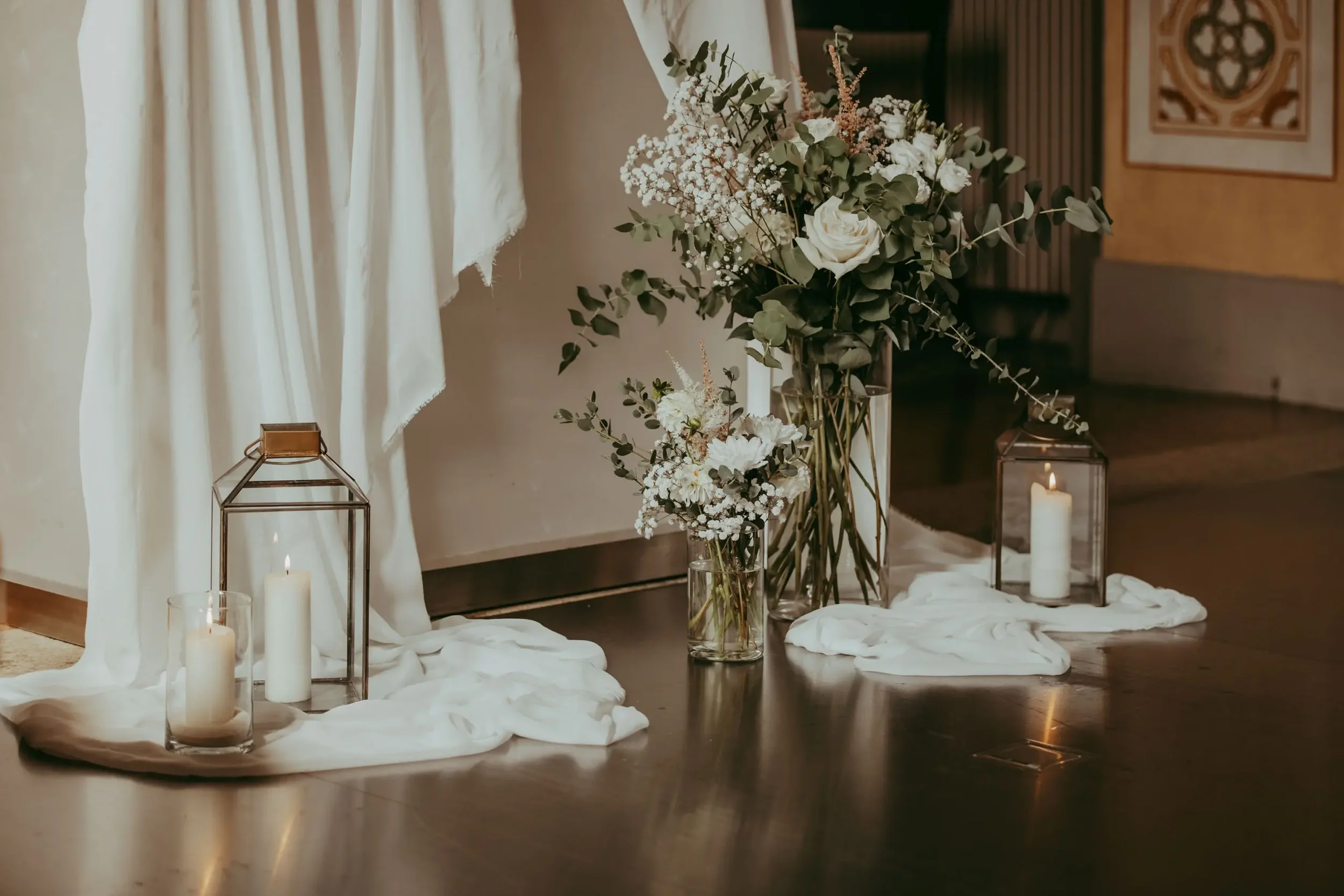 Blumenstrauß mit weißen Rosen, Eukalyptus und kleinen weißen Blumen in Glasvasen, umgeben von Kerzen in Glasgefäßen, auf einem weißen Tuch, vor einem beigeweißen Vorhang und einer Wand mit Dekoration.