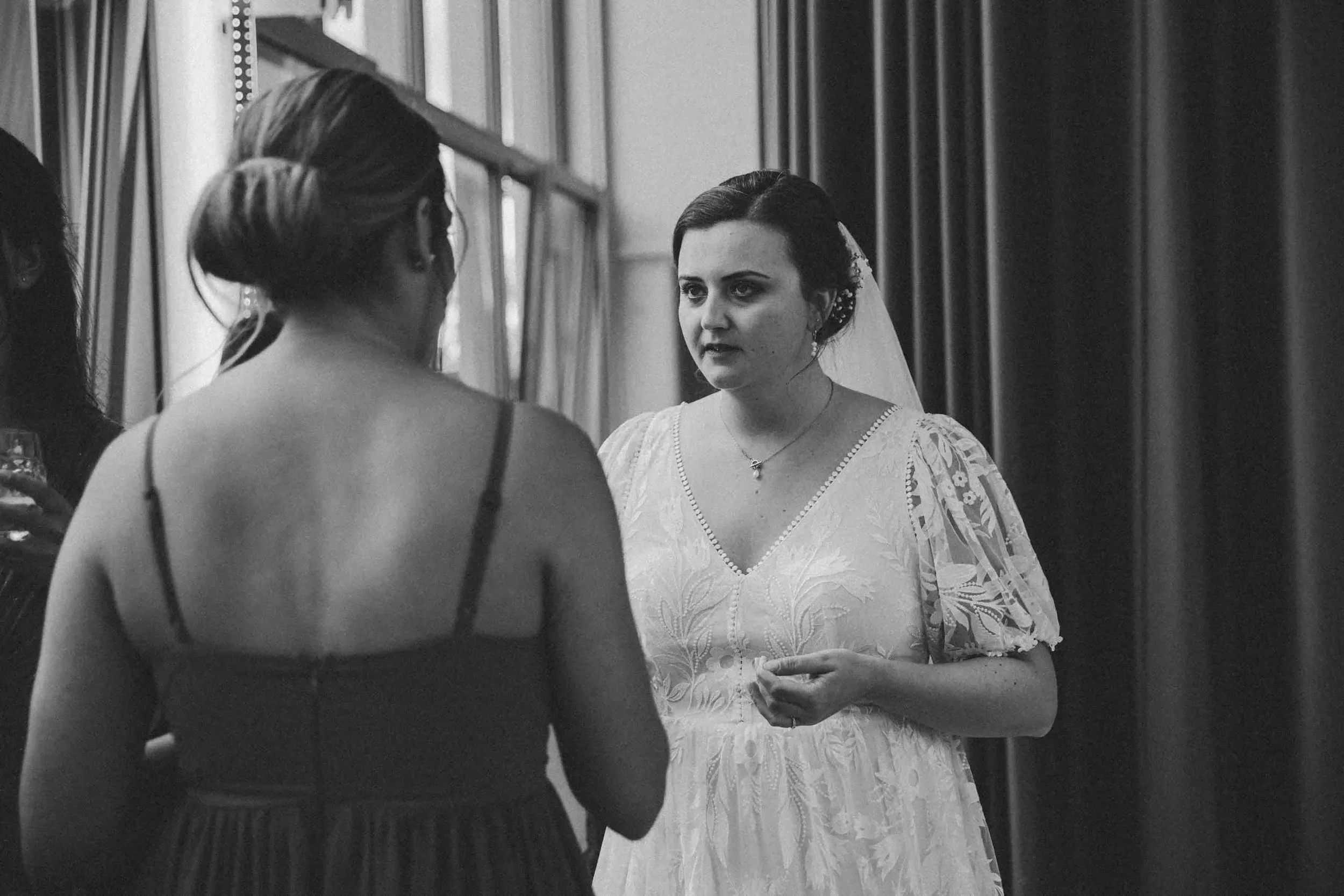 Schwarz-Weiß-Foto von zwei Frauen, die sich bei einer Hochzeit unterhalten, eine trägt ein weißes Hochzeitskleid mit Blumenmuster und eine Haarnadel, die andere trägt ein dunkleres Kleid.