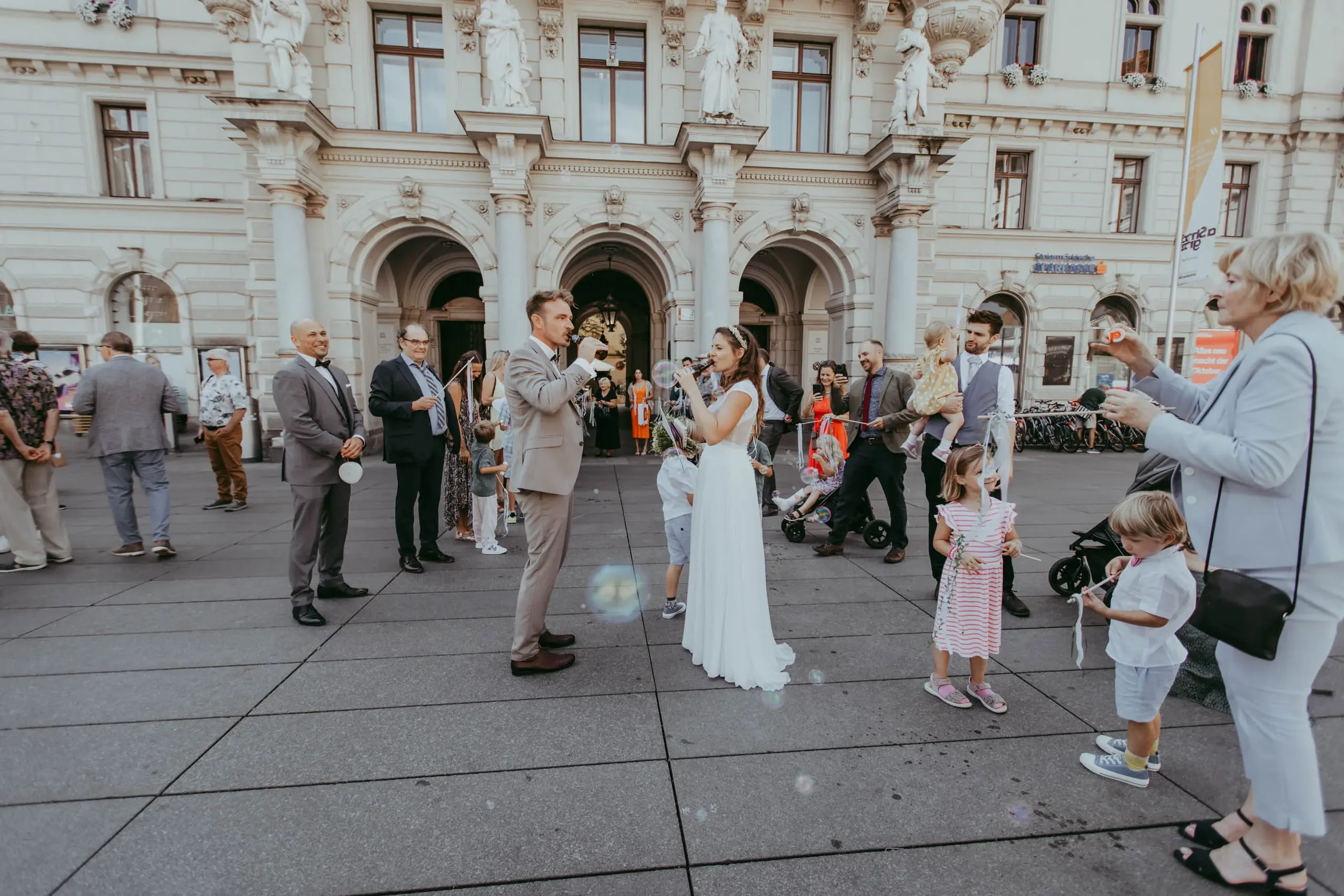 Menschen bei einer Hochzeit im öffentlichen Raum, einige sind Kinder, andere Erwachsene, im Hintergrund ein historisches Gebäude.