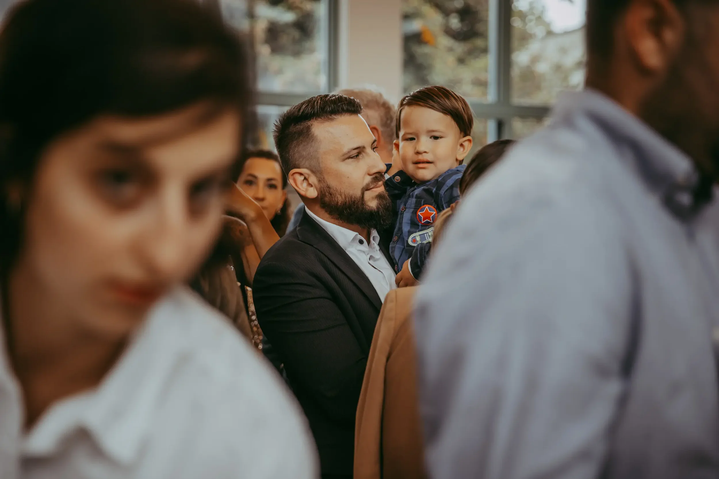 Menschen in einem Raum, darunter ein Mann mit Bart, der ein Kind auf dem Arm hält, und weitere Personen im Hintergrund, vermutlich bei einer Veranstaltung oder Versammlung.