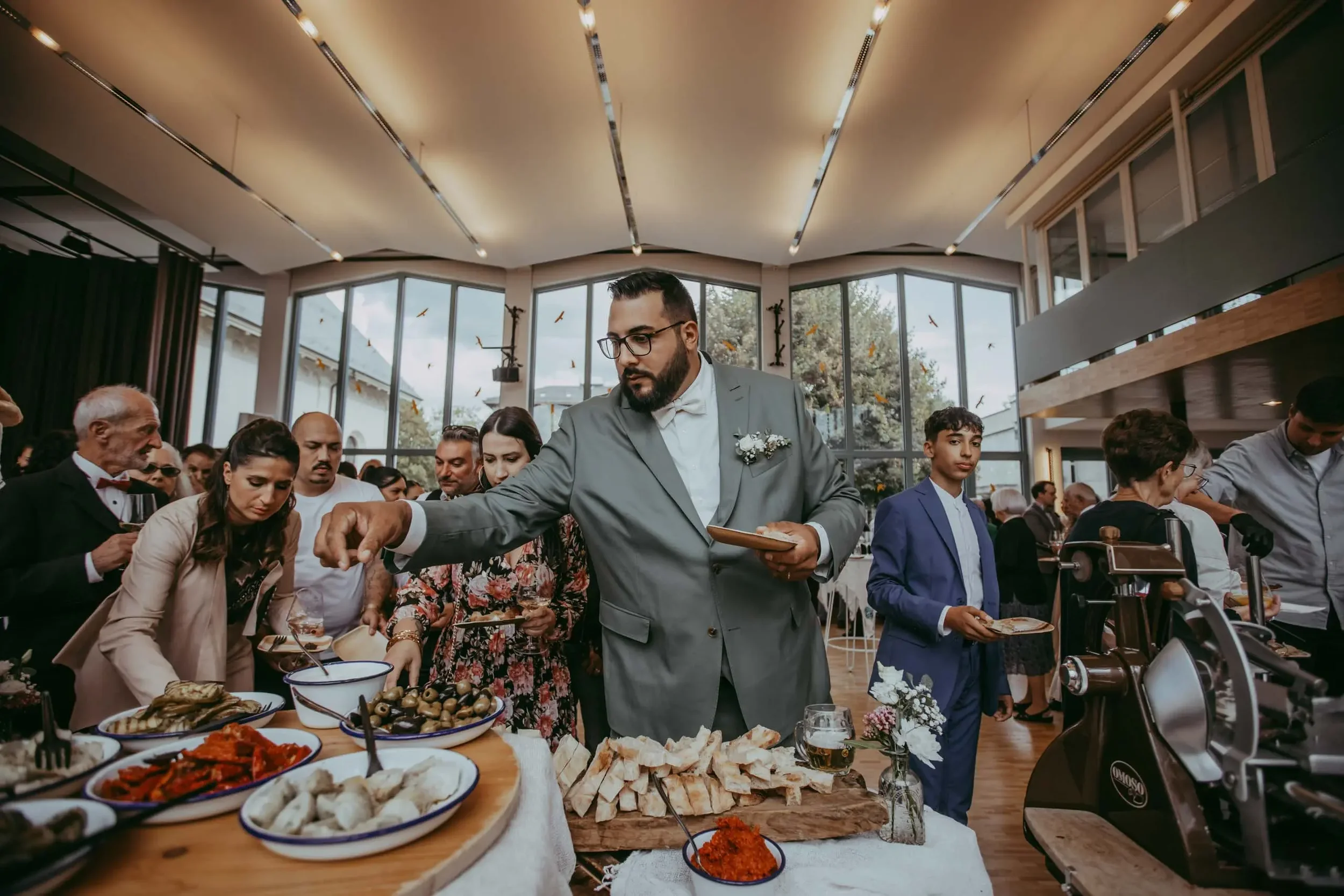 Menschen bei einer Veranstaltung, die Essen von einem Buffet nehmen, in einem modernen Raum mit großen Fenstern und natürlichem Licht.