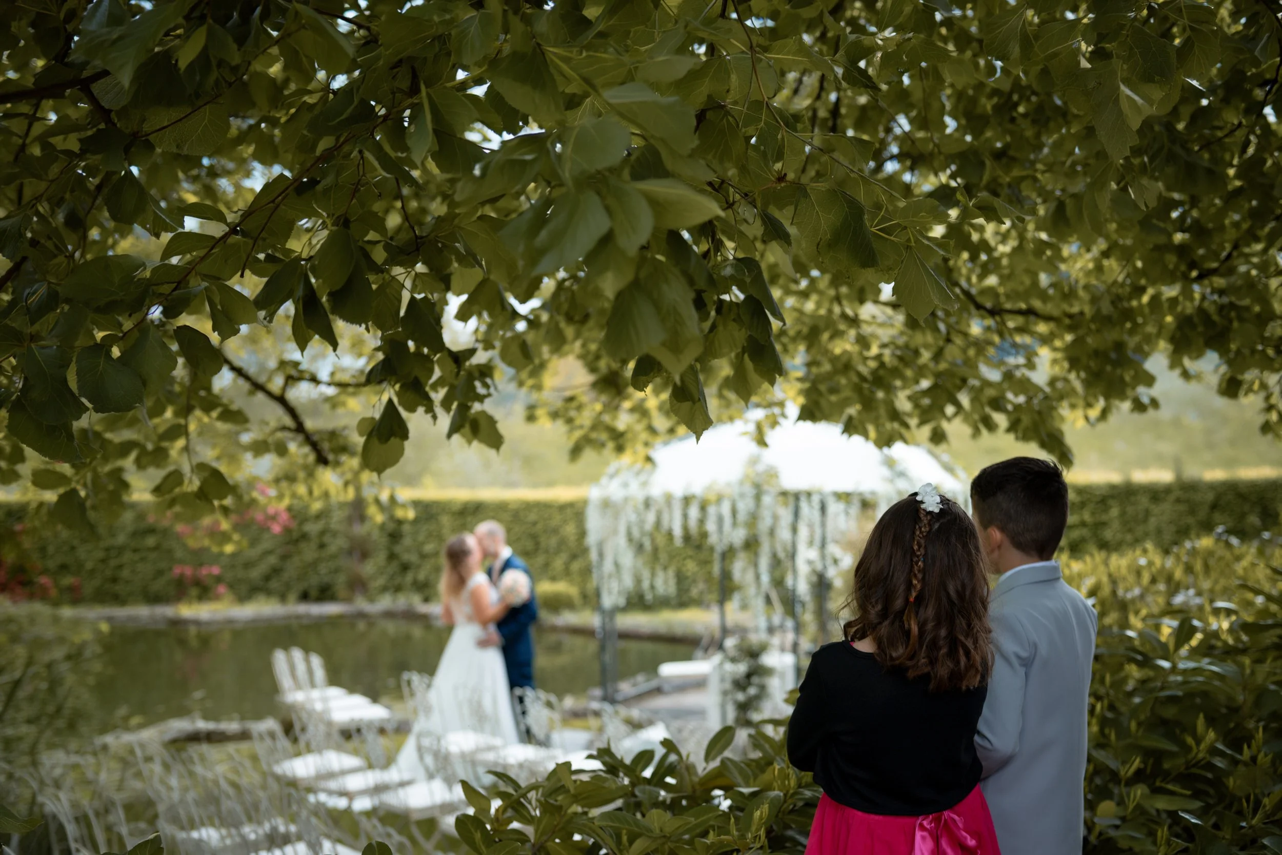 Ein Bild von einer Hochzeit im Freien, gesehen durch Blätter eines Baumes. Im Hintergrund sind ein Brautpaar, eine Brautjungfer und ein Bräutigam zu erkennen, die sich gegenüberstehen. Weitere Personen sind teilweise sichtbar, es gibt auch weiße Stüh