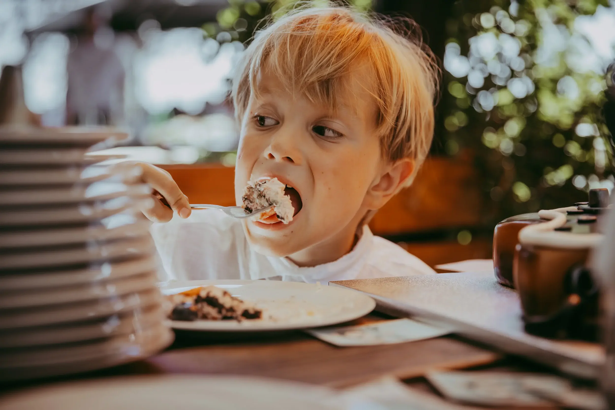 Junge mit roten Haaren isst Kuchen in einem Restaurant. Im Hintergrund sind Pflanzen und bunte Lichter, im Vordergrund ein Teller mit Kuchen und eine Reihe von Torten.