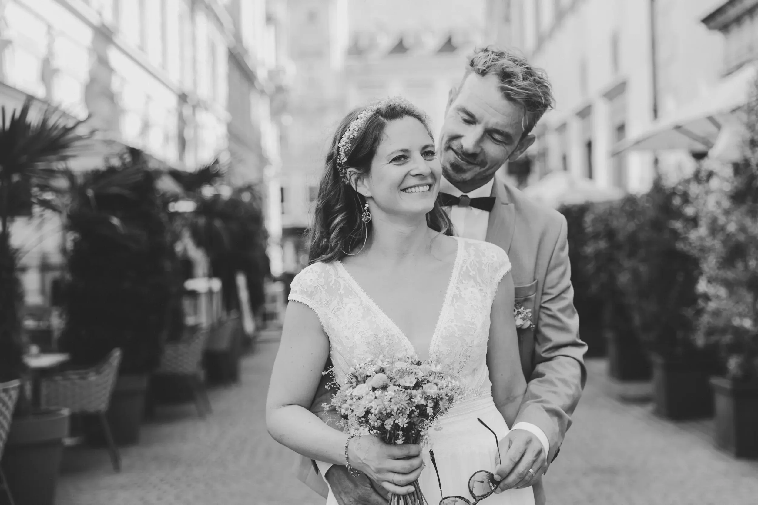 Ein lachendes Brautpaar, die Braut trägt ein weißes Kleid mit Spitze und eine Blumenkrone, der Bräutigam trägt einen Anzug mit Fliege, steht im Freien in einer urbanen Umgebung.