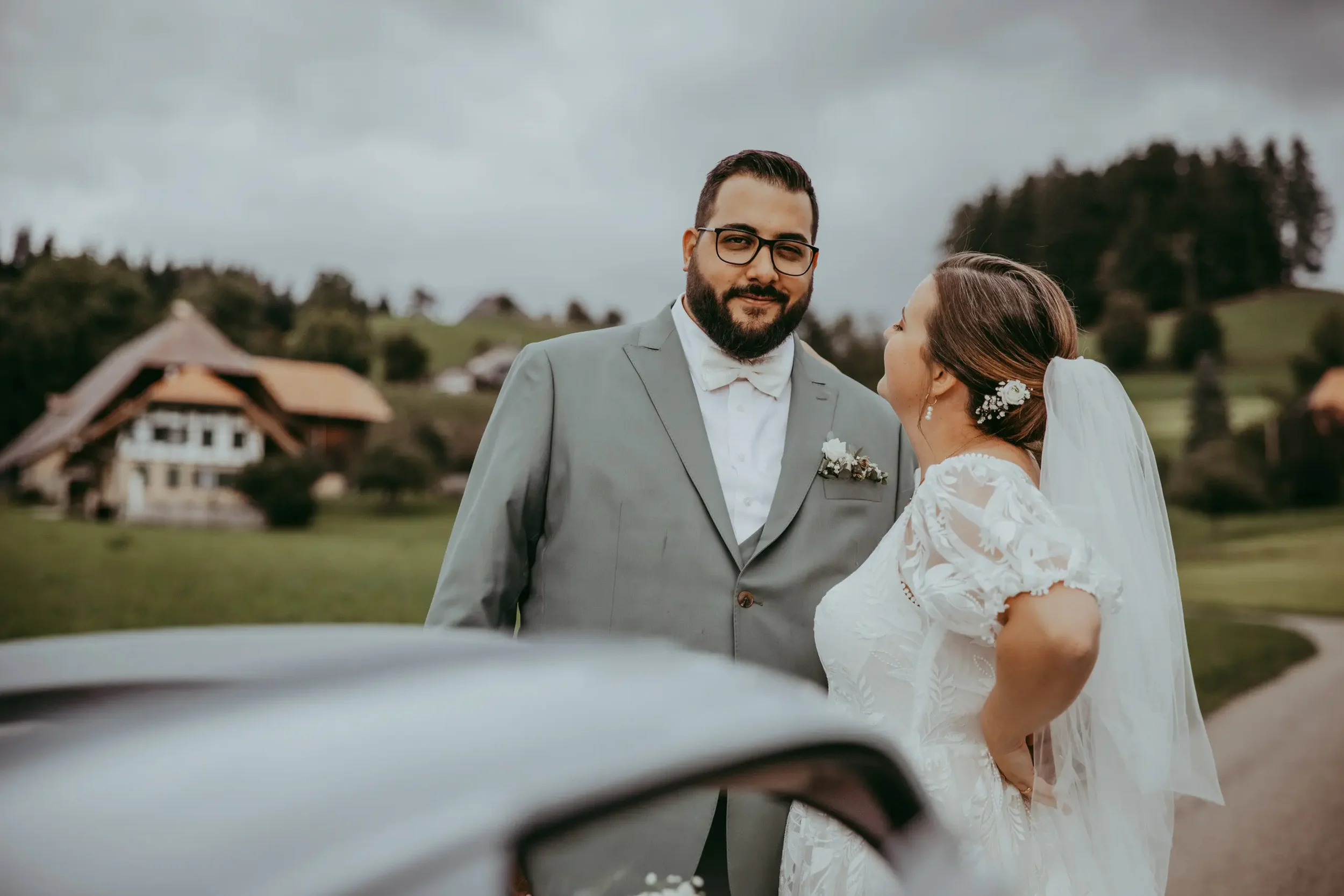 Ein Mann in einem grauen Anzug und eine Frau in einem weißen Brautkleid stehen sich bei einer Hochzeit im Freien gegenüber, während der Himmel bewölkt ist.